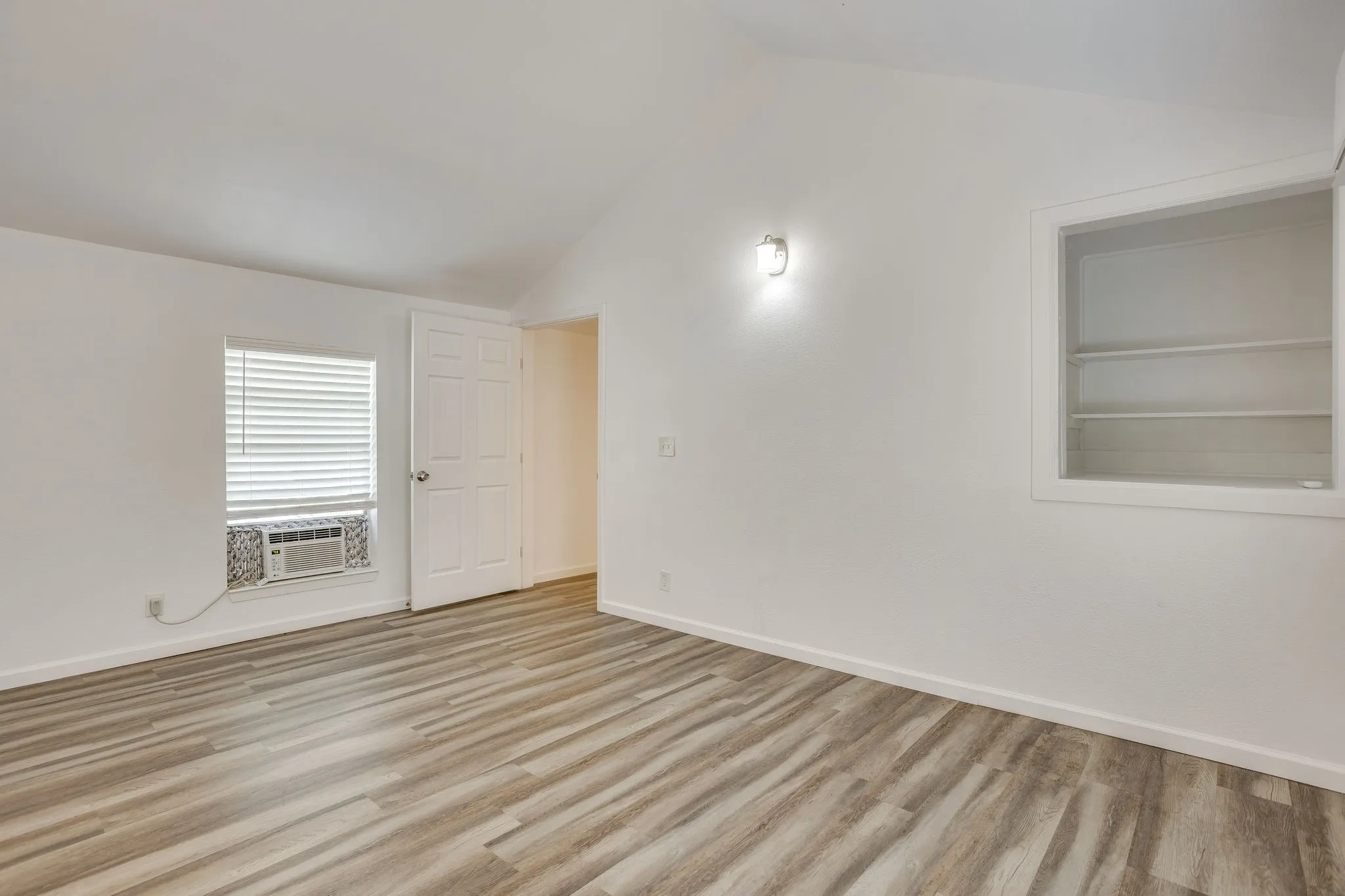 Vaulted ceiling with a wall-mounted light fixture, wood-finish flooring, built-in shelving, and a window with blinds