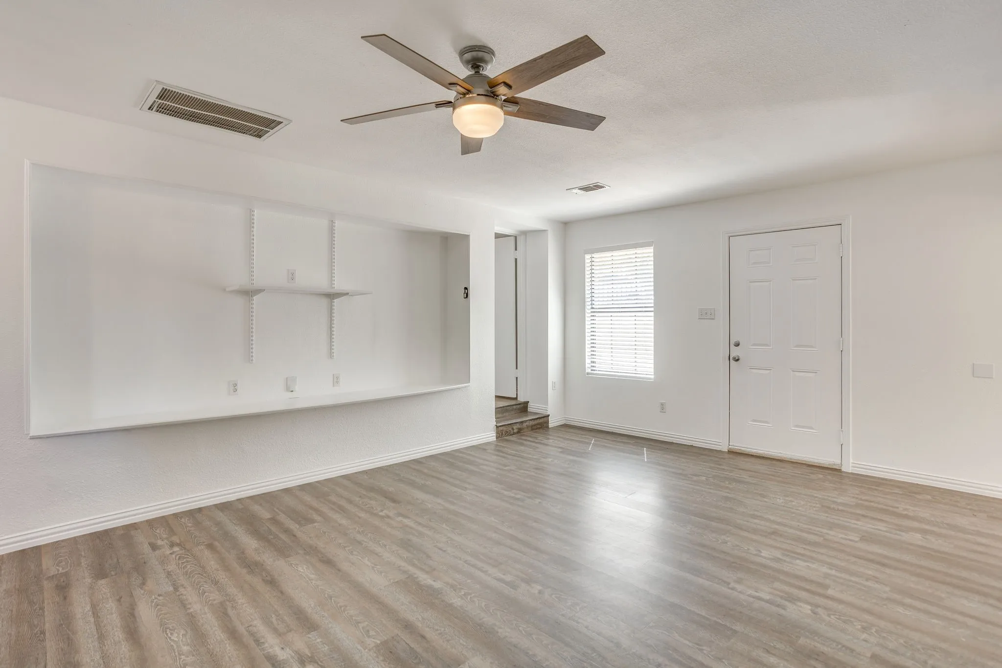 Wood-finish flooring throughout, ceiling fan with integrated lighting, built-in shelving, white paneled door, and window with blinds