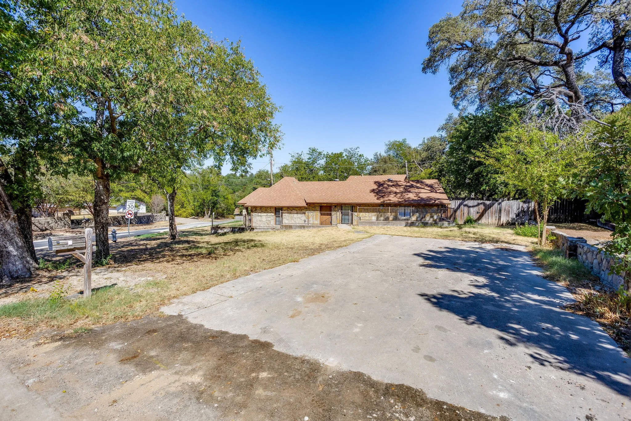 Stone facade residence with a brown shingled roof, set back from a paved driveway, featuring mature trees on the property