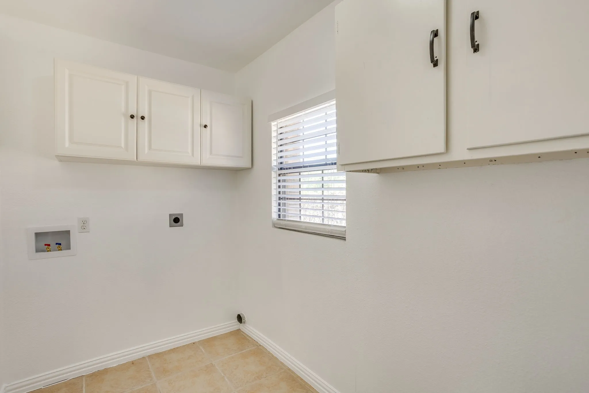 Tile flooring, white wall-mounted cabinetry, single window with blinds, and appliance hookups