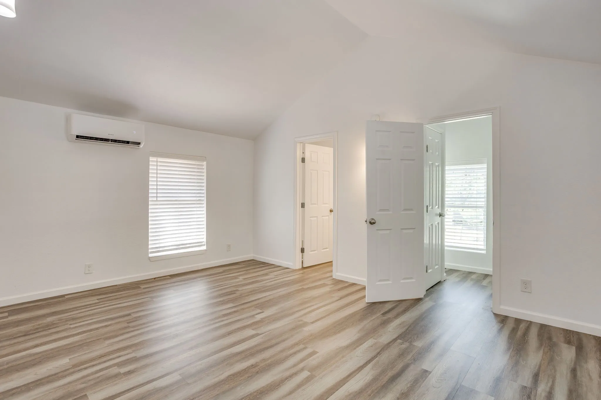 Vaulted ceiling with a ceiling-mounted lighting fixture, wood-finish flooring, white paneled doors, and a split-system air conditioning unit