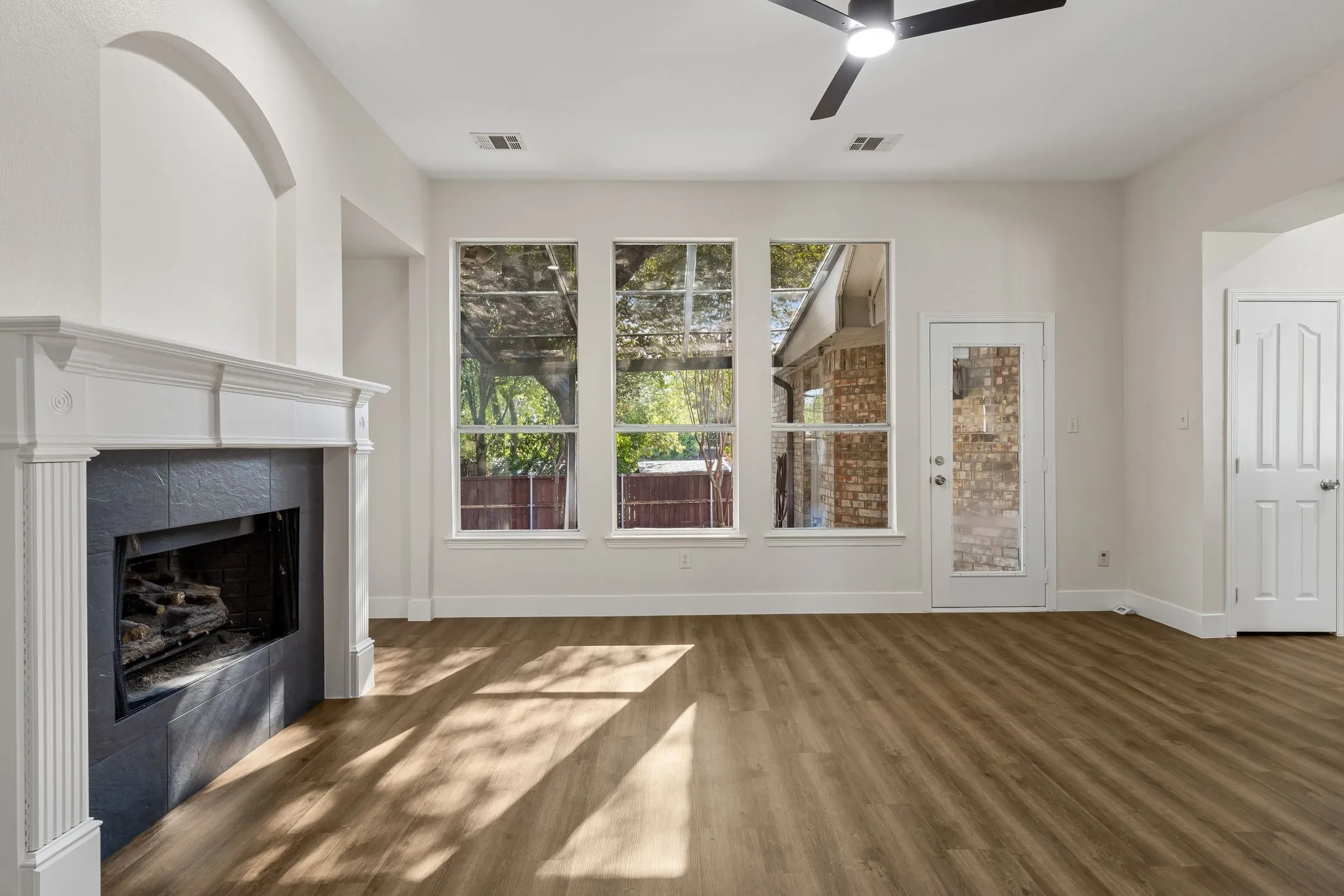 White fireplace with a dark tile surround, wood-finish flooring, three large windows, and a ceiling fan