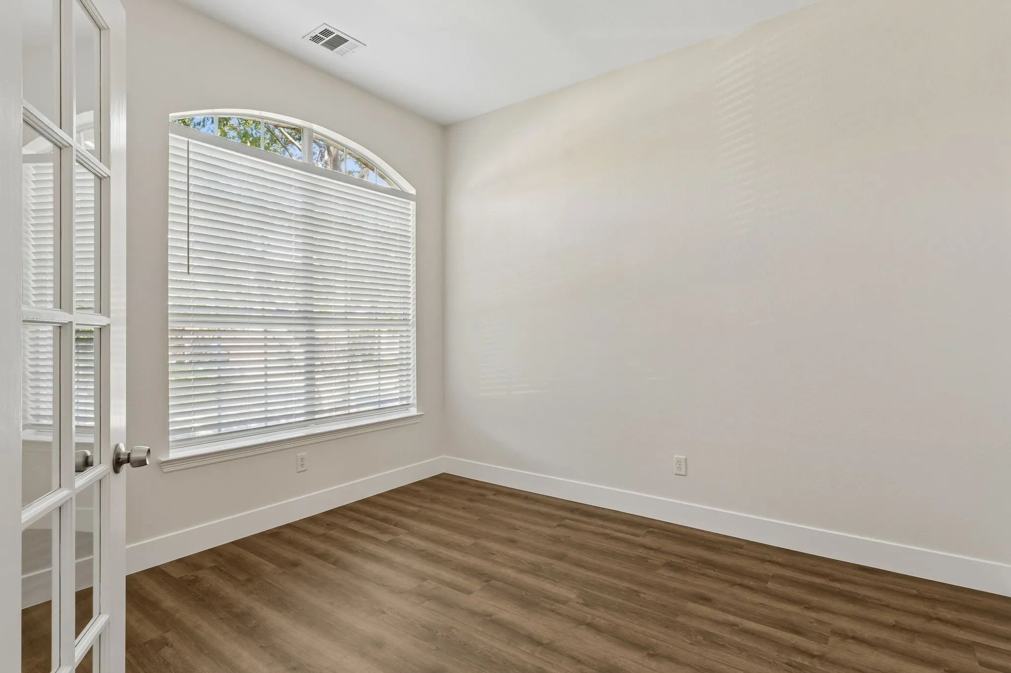 Arched window with blinds, wood-finish flooring, white trim, and a white paneled door