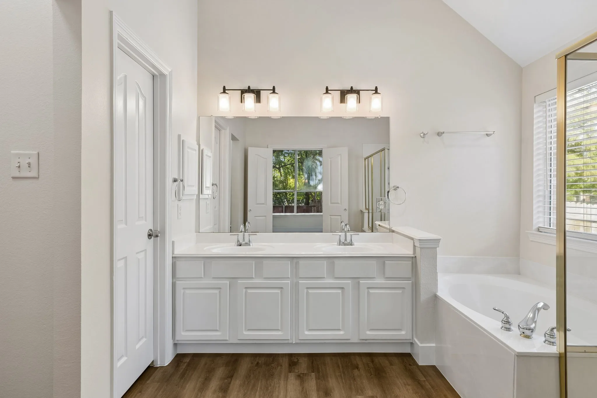 Dual vanity with white cabinetry, wood-finish flooring, built-in bathtub, and overhead lighting