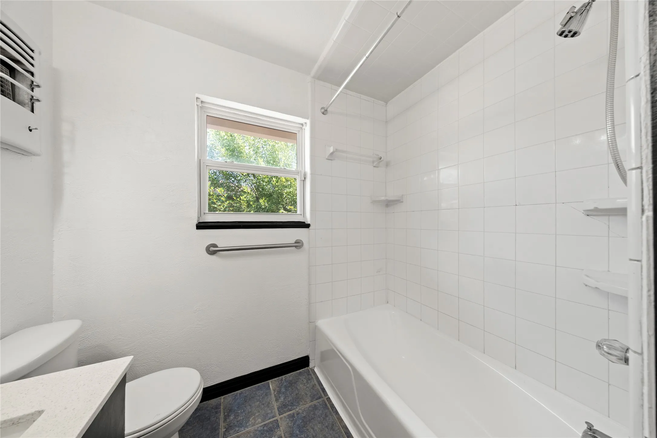 Bathtub with white tile surround, a window with a privacy blind, and a toilet with a white countertop