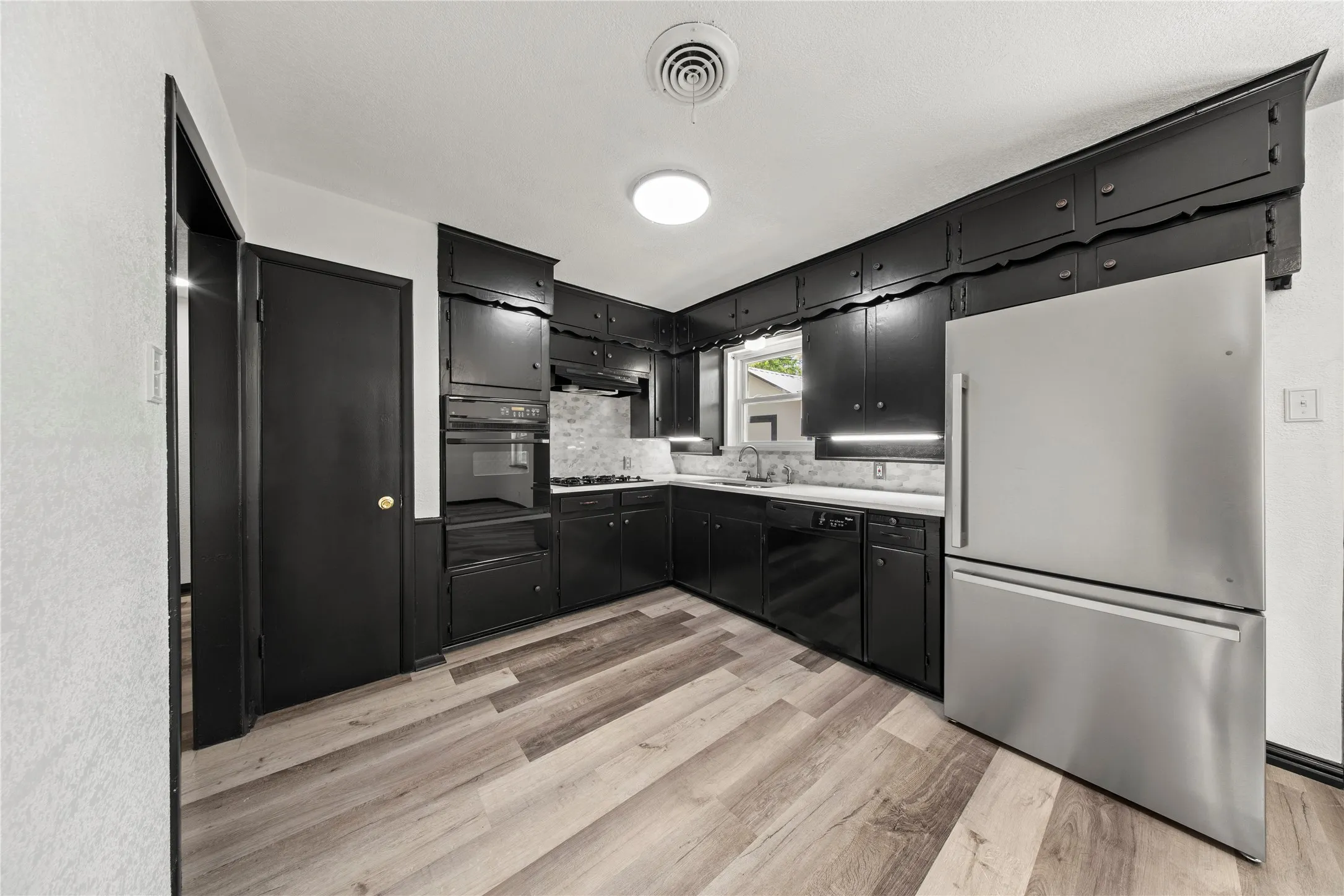 Kitchen featuring wood-finish flooring, black cabinetry, white countertops, built-in oven, and a stainless steel refrigerator
