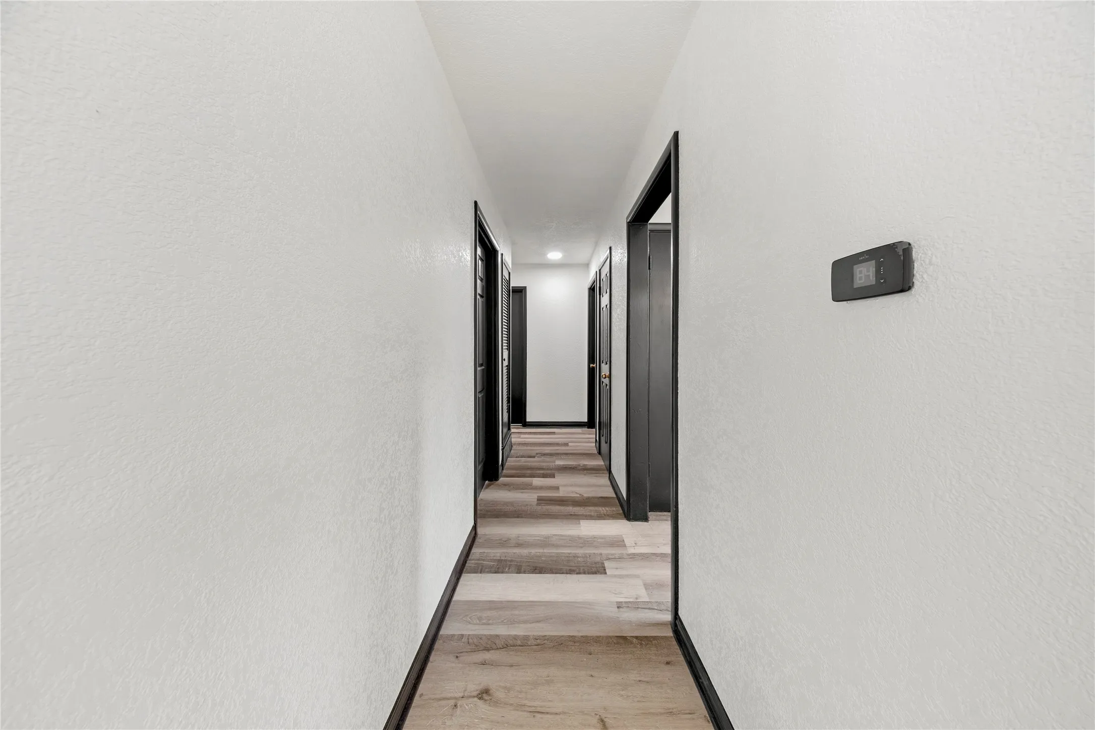 Hallway featuring wood-finish flooring, white walls, dark door frames, and a wall-mounted thermostat