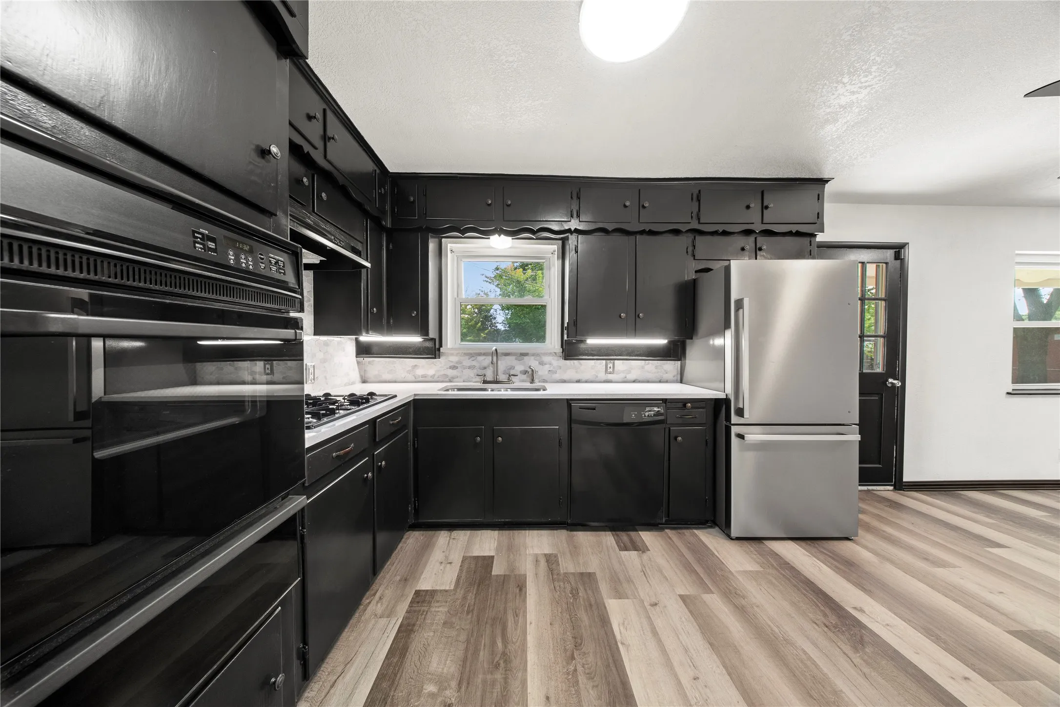 Kitchen featuring black cabinetry, stainless steel appliances, white countertops, a mosaic tile backsplash, and wood-finish flooring