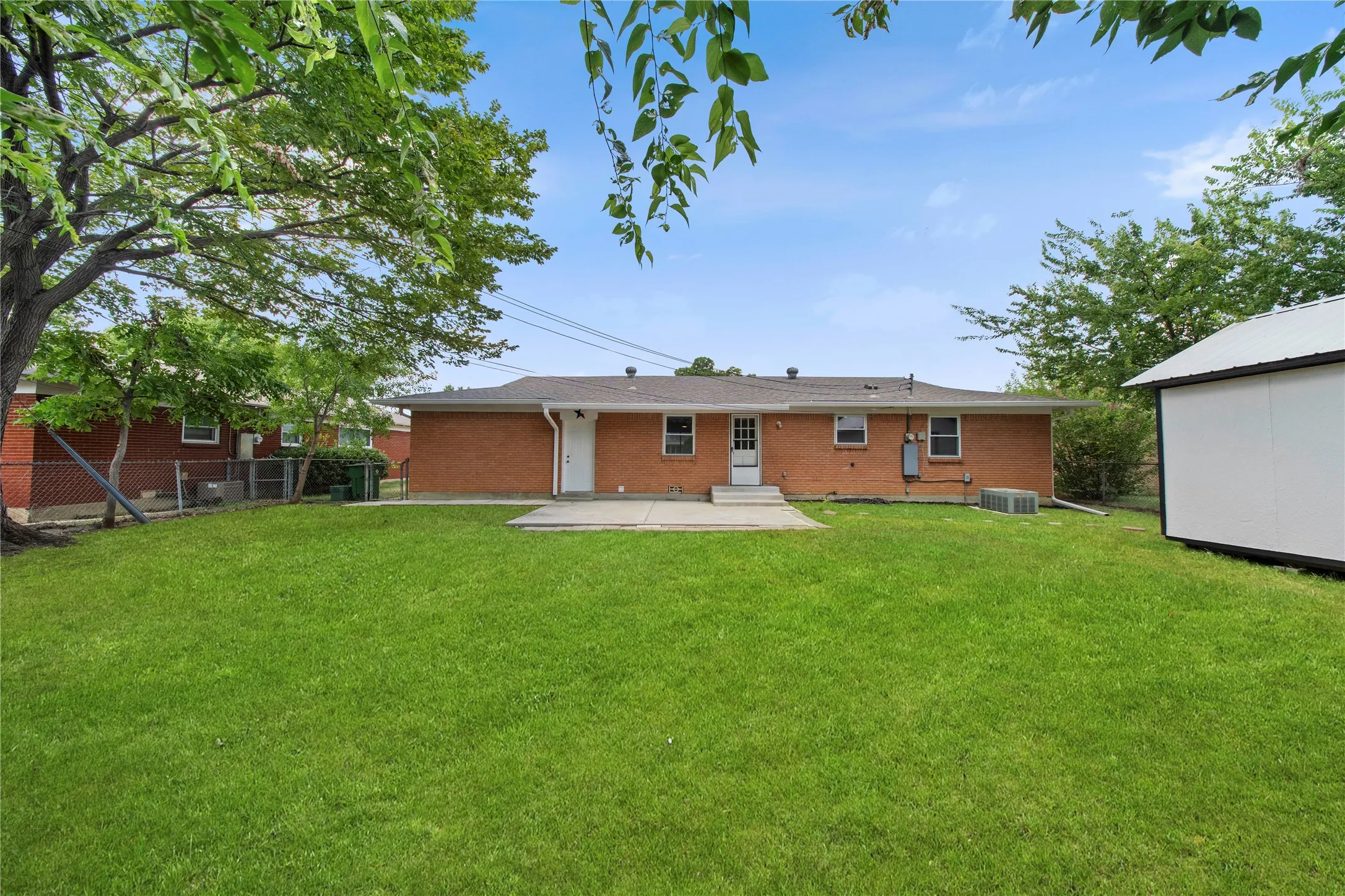 Expansive rear lawn, brick exterior, concrete patio, white storage shed, and mature trees