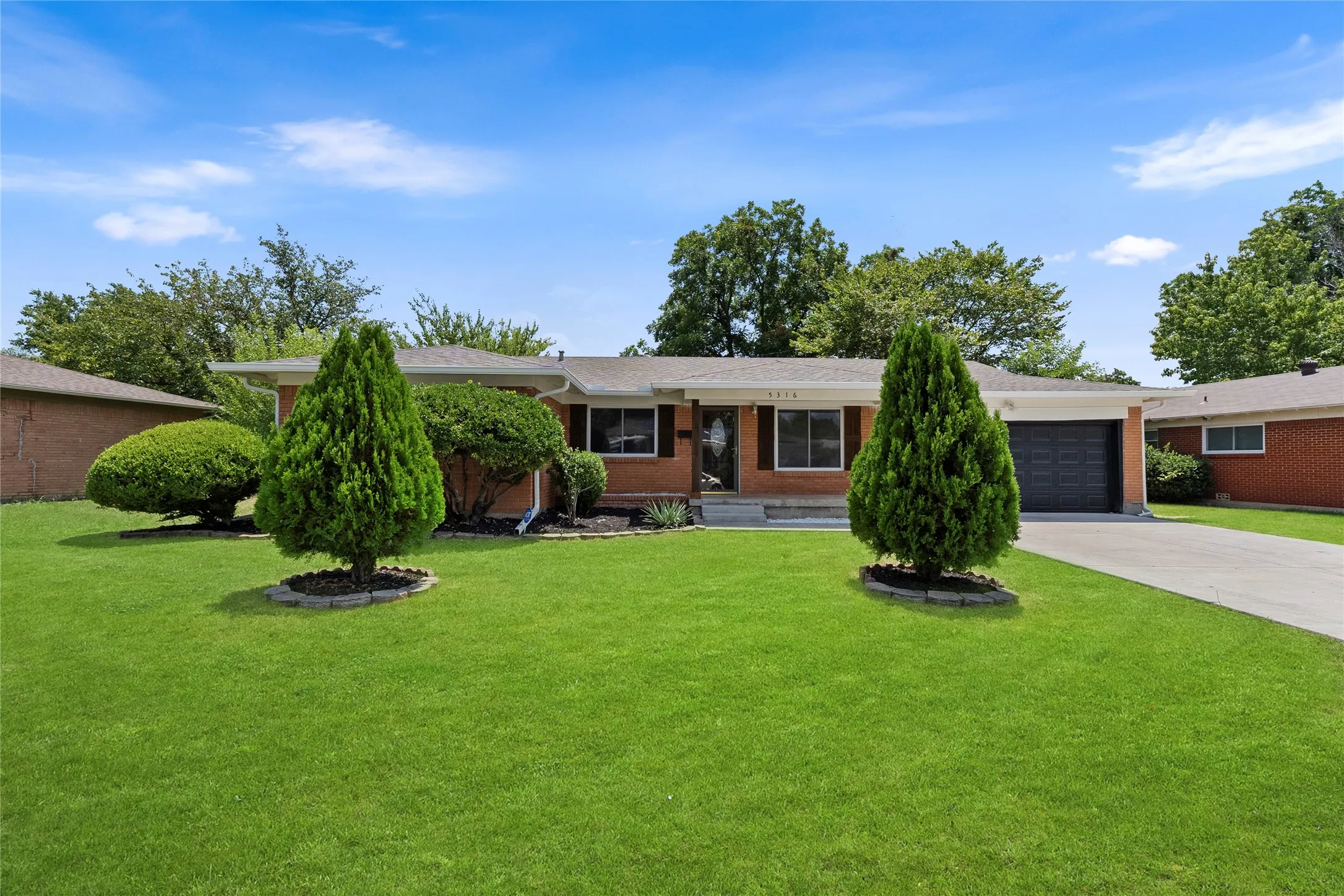 Brick exterior with a dark garage door, concrete driveway, front lawn, and established landscaping