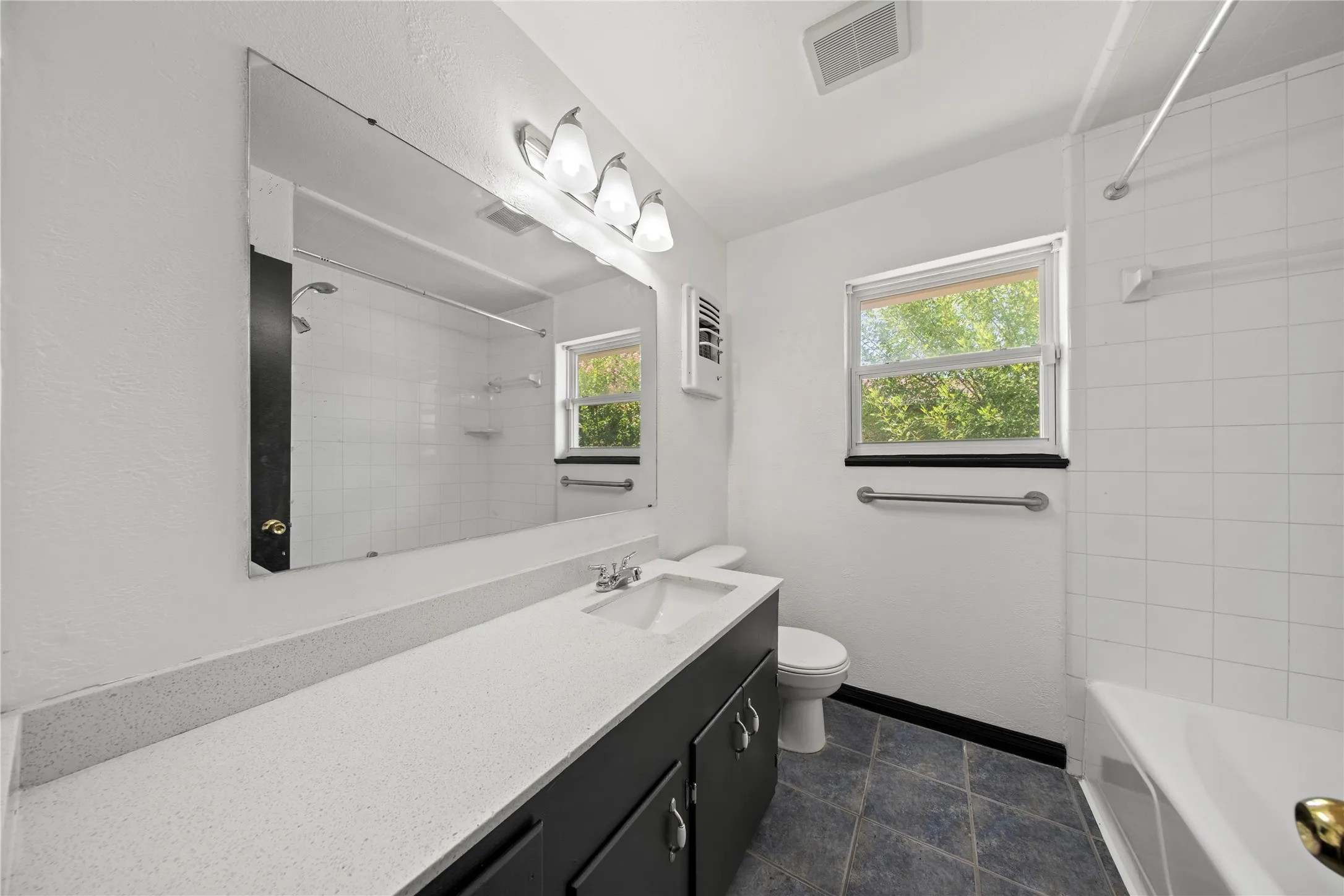 Vanity with an integrated sink, expansive wall mirror, window with natural light, bathtub with tile surround, and patterned floor tiling