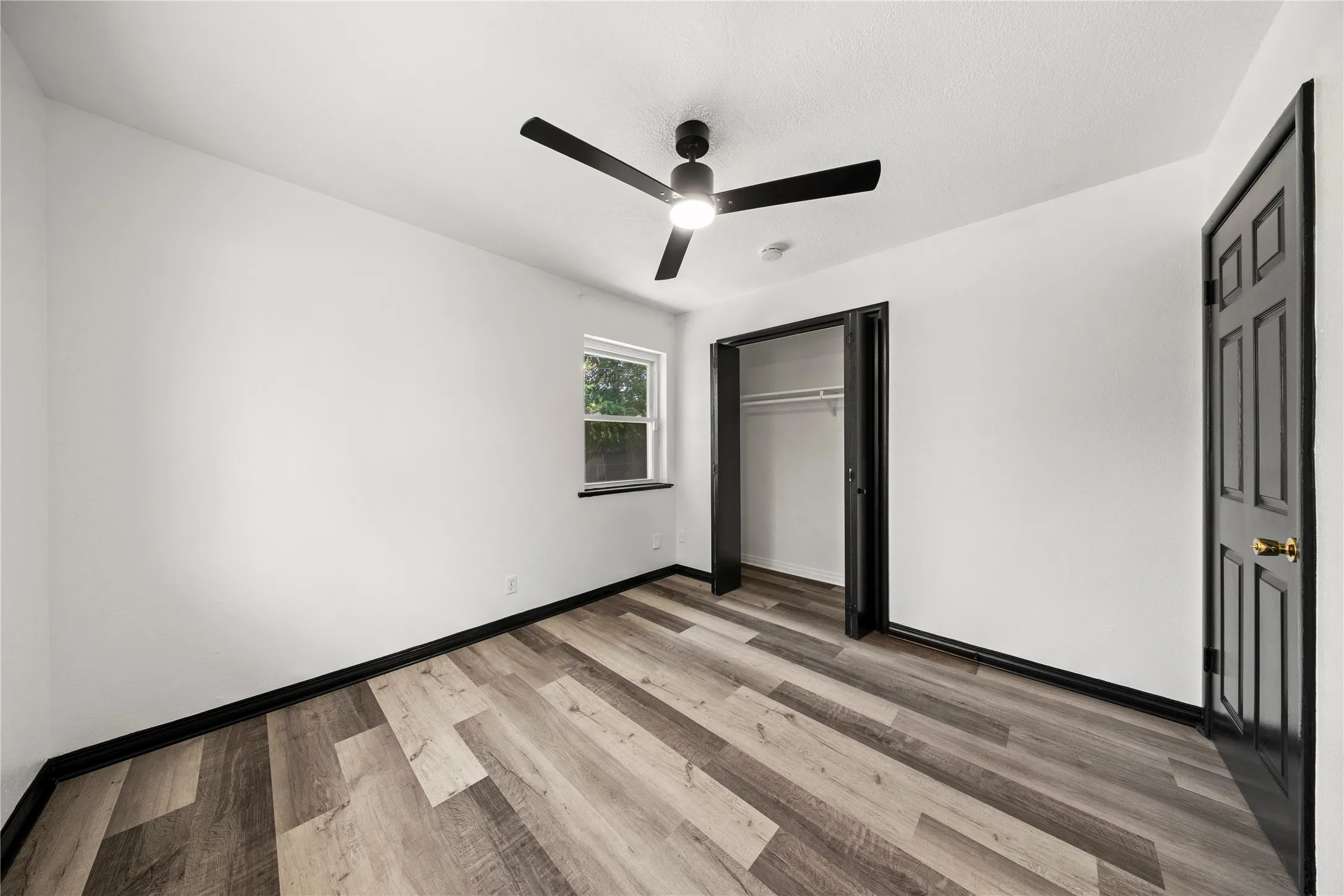 Wood-finish flooring, white walls, black trim, window with exterior view, and ceiling fan with integrated lighting