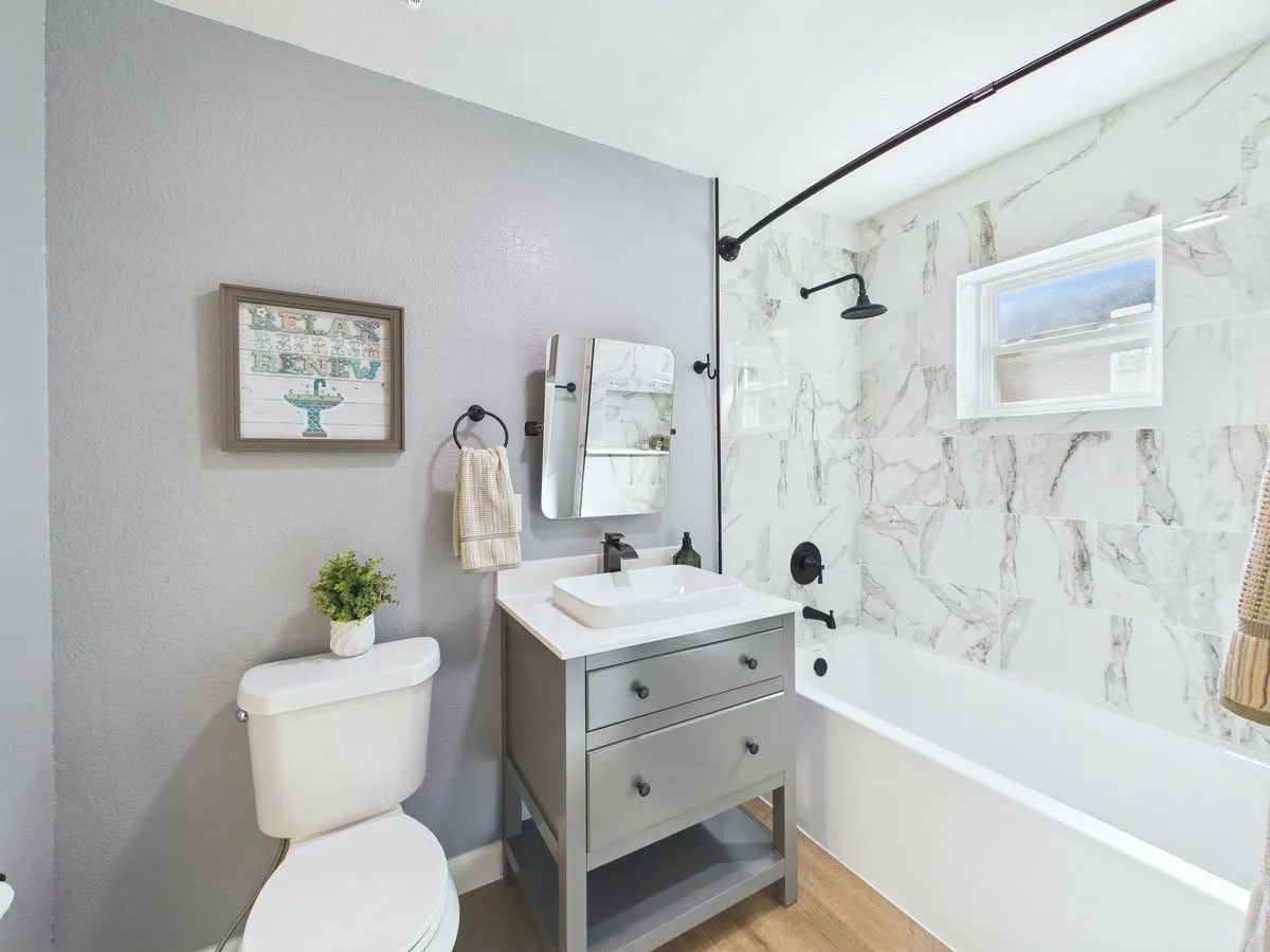 Wall-mounted shower head with matte black fixtures, white rectangular bathtub, wood-finish flooring, vanity with integrated vessel sink, and large format wall tile