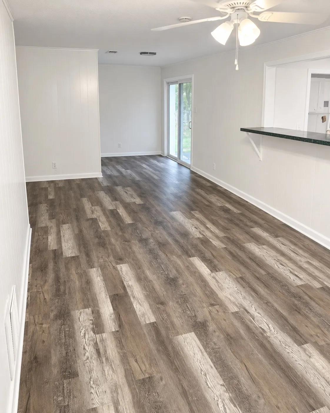 Wood-finish plank flooring, ceiling fan with integrated lighting, sliding glass door, and a pass-through counter with a dark countertop