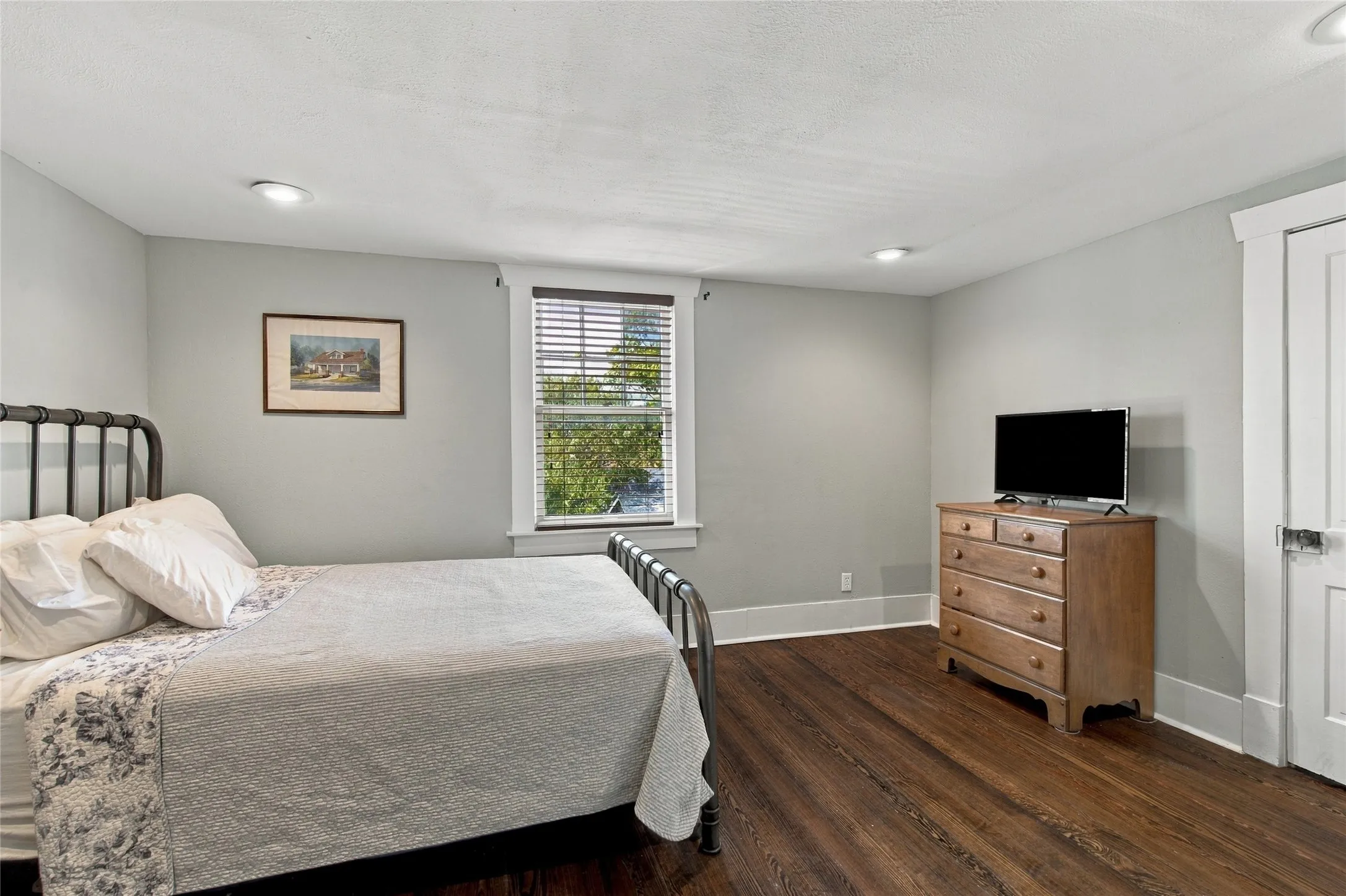 Rich wood-finish flooring, recessed ceiling lighting, a window with horizontal blinds, and a paneled door with a lever handle
