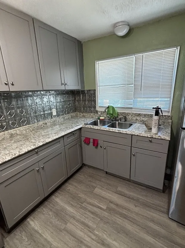 Gray cabinetry, patterned backsplash, light-toned countertops, double basin sink, and wood-finish flooring