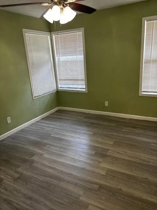 Wood-finish flooring with white trim, two windows with blinds, an additional window with blinds, and a ceiling fan with integrated lighting