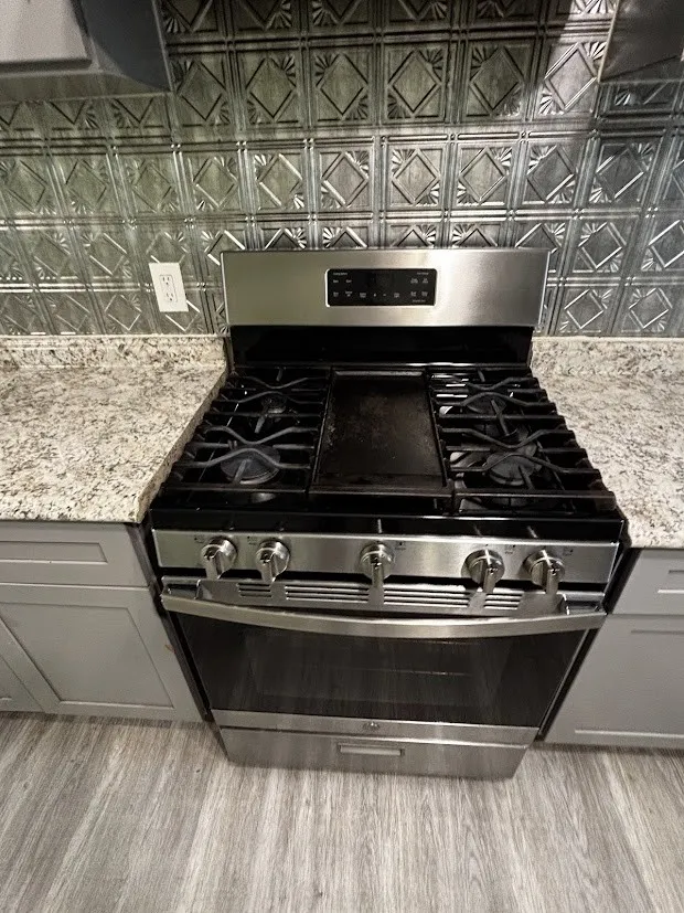 Stainless steel range with a built-in griddle, granite-style countertops, decorative metal backsplash, wood-finish flooring, and shaker-style cabinetry