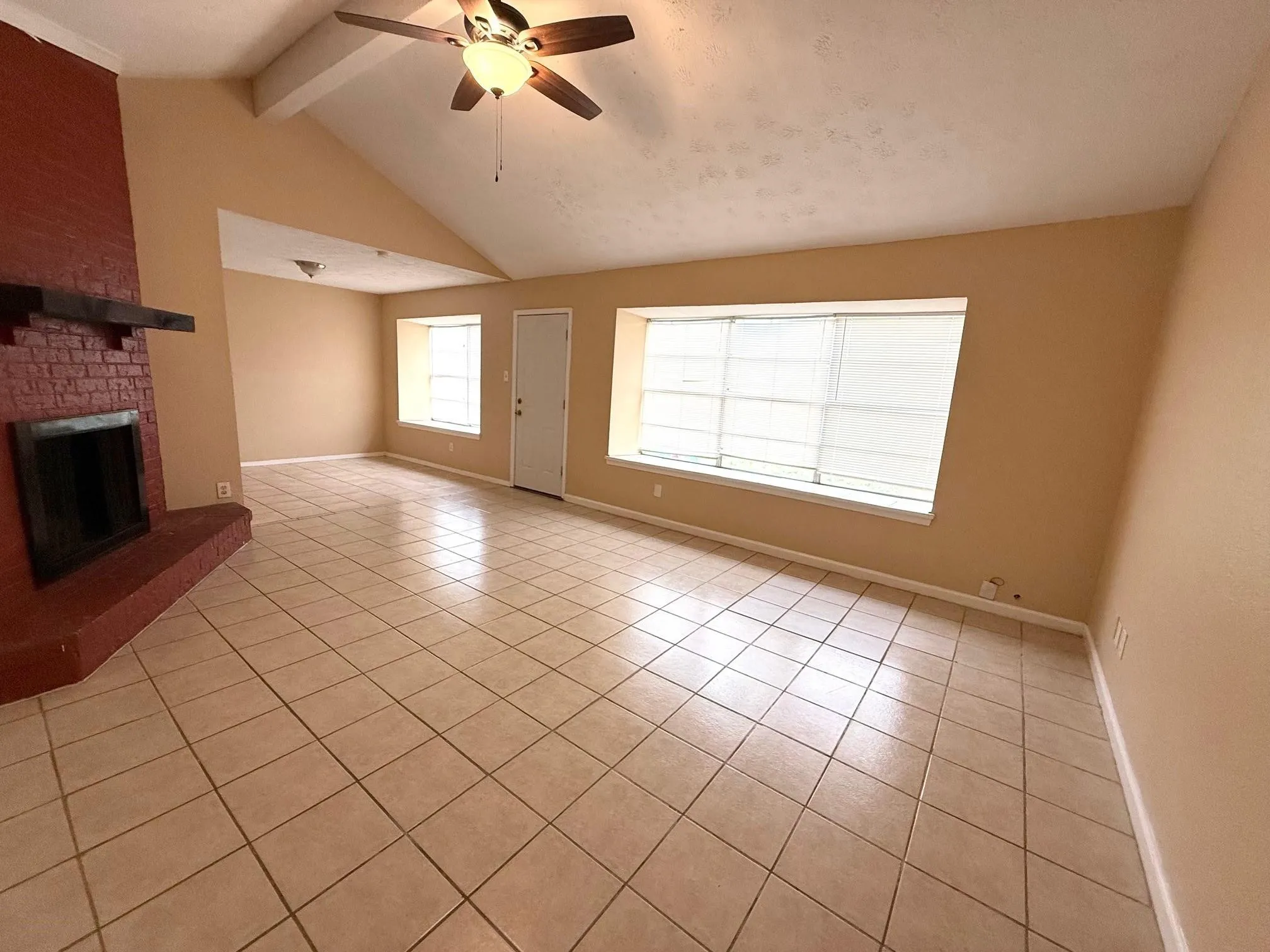 Vaulted ceilings with exposed beam, ceramic tile flooring, brick fireplace with wood mantel, ceiling fan with light fixture, and window with blinds