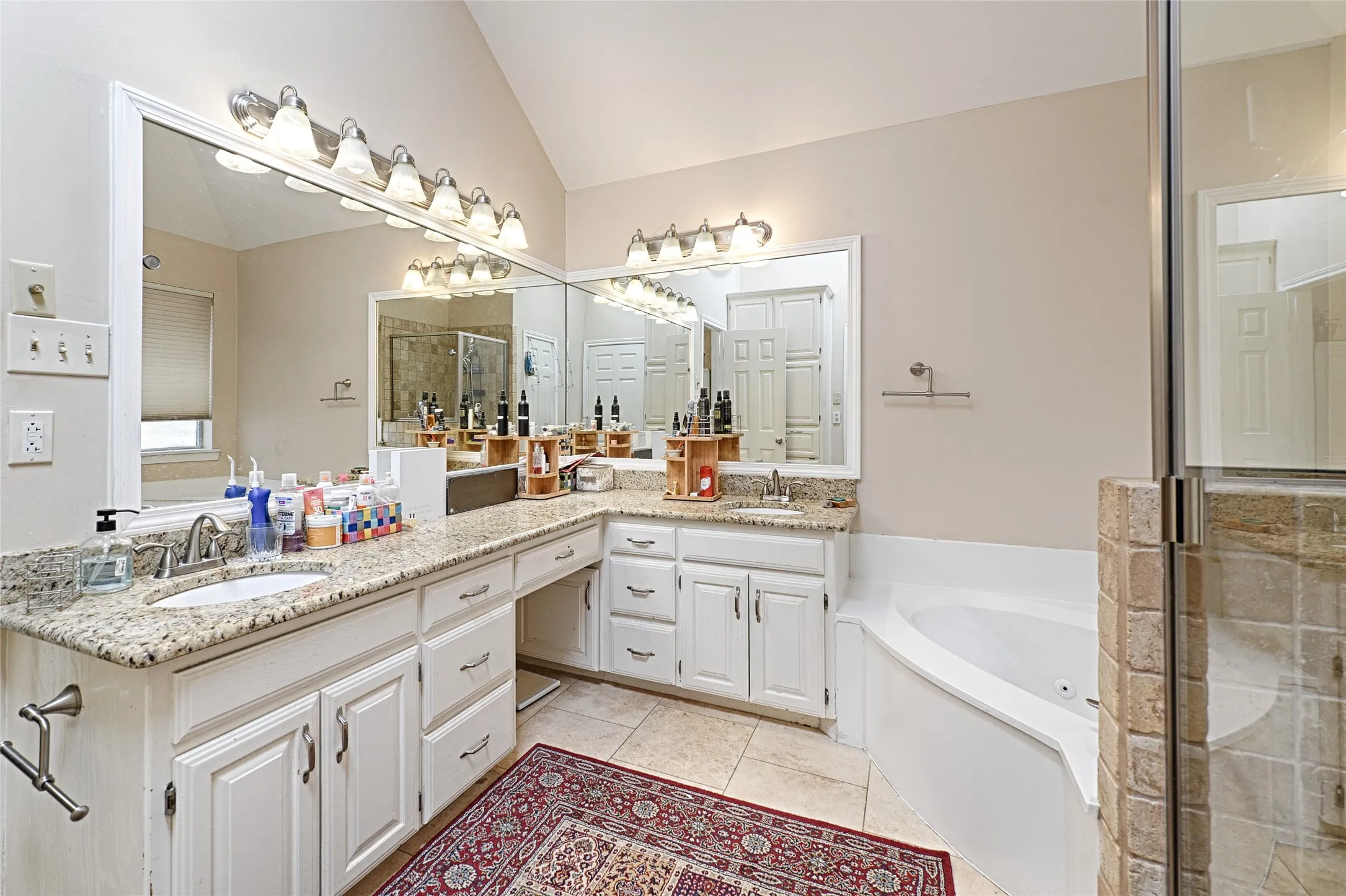 Dual vanities with granite countertops, integrated mirrors, white cabinetry, a corner soaking tub, and a glass-enclosed shower with tile surround