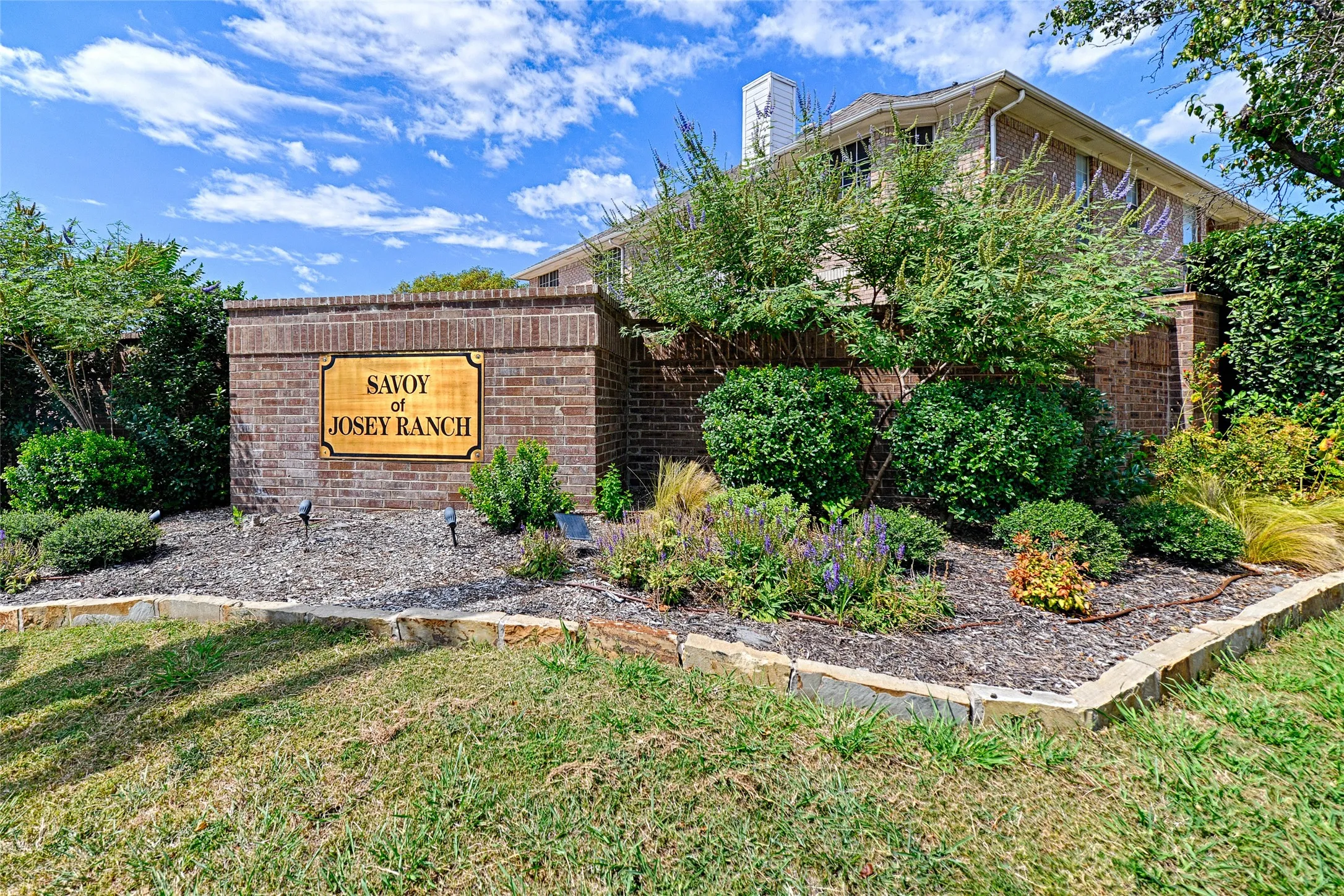 Brick-clad entry monument with a carved wooden sign, bordered by a stone-edged planting bed, lush landscaping, and mature trees
