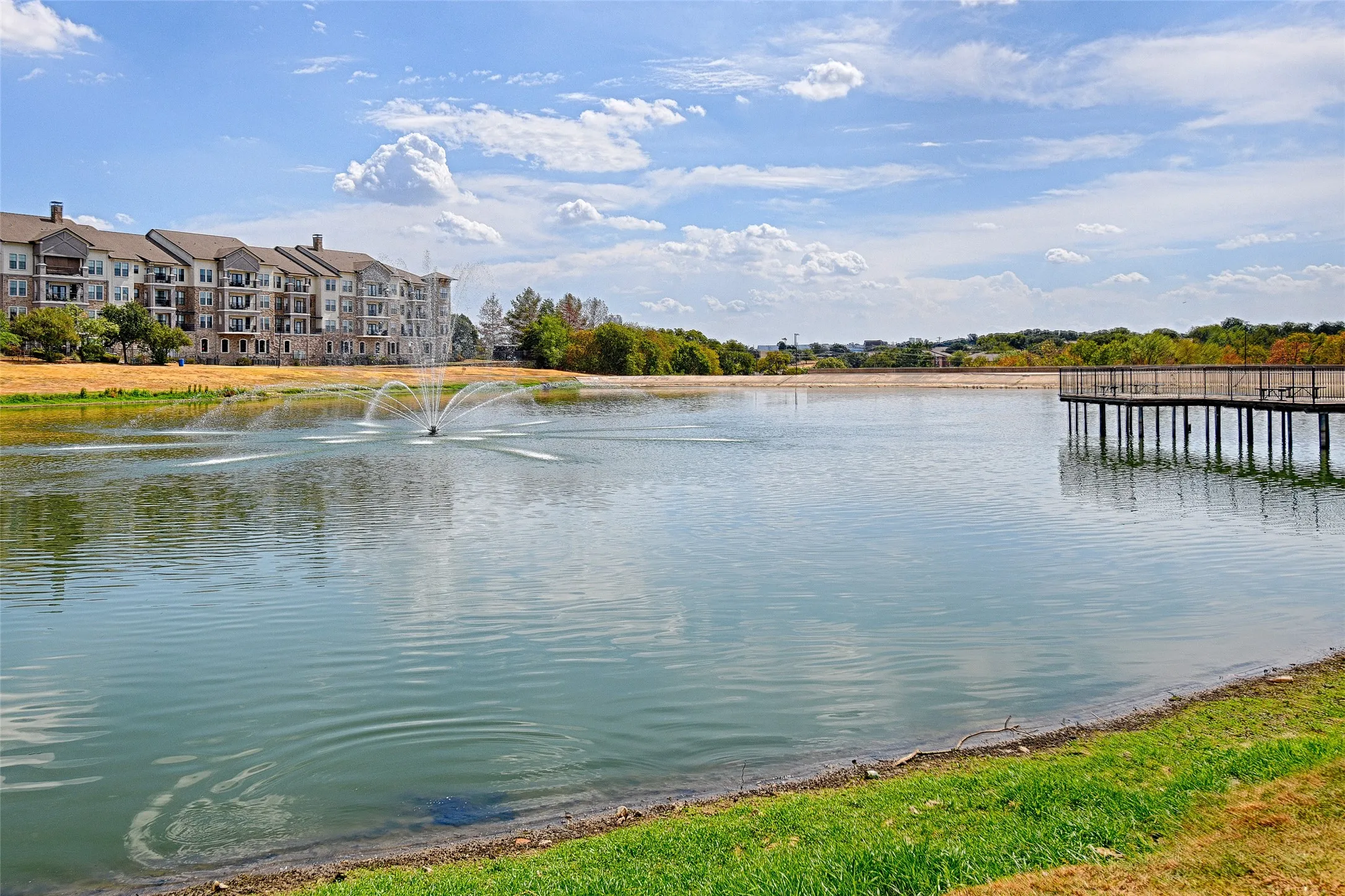Water feature with fountain, adjacent at Josey Ranch Park Senior residential building, and a wood-decked pier extending over the water