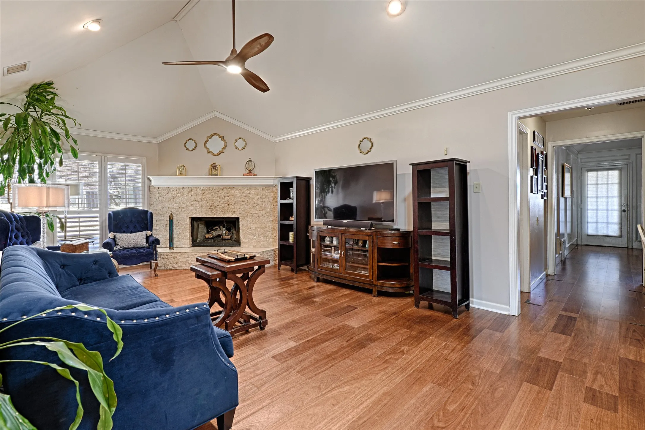 Vaulted ceiling with a ceiling fan, wood-finish flooring, stone fireplace, and crown molding