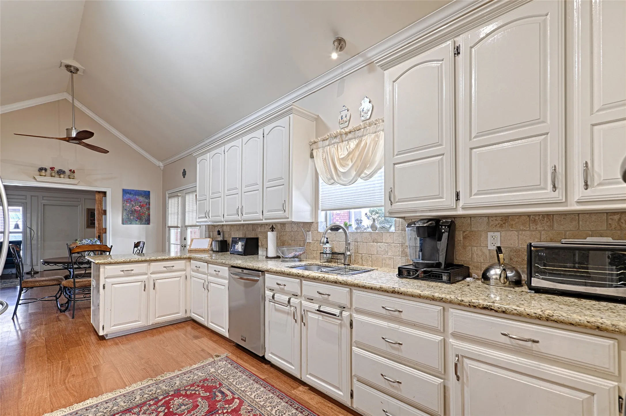 Vaulted ceiling with a ceiling fan, white cabinetry with crown molding, granite-finish countertops, stainless steel dishwasher, and wood-finish flooring