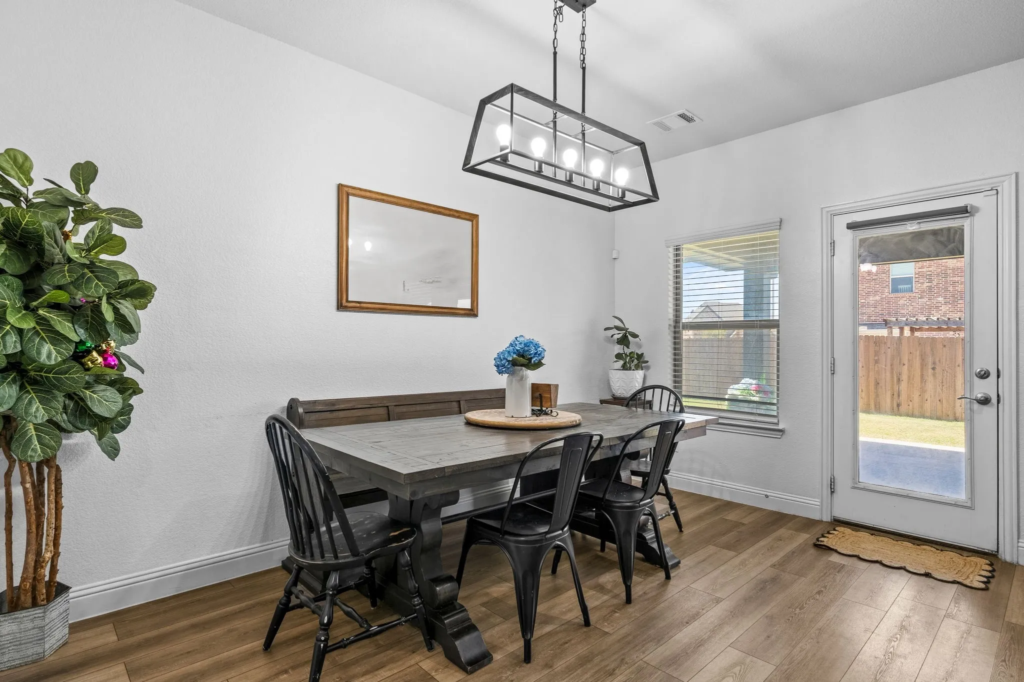 Open-concept dining area featuring wood-finish flooring, a modern linear chandelier, a glass-panel exterior door, and a privacy-fenced backyard