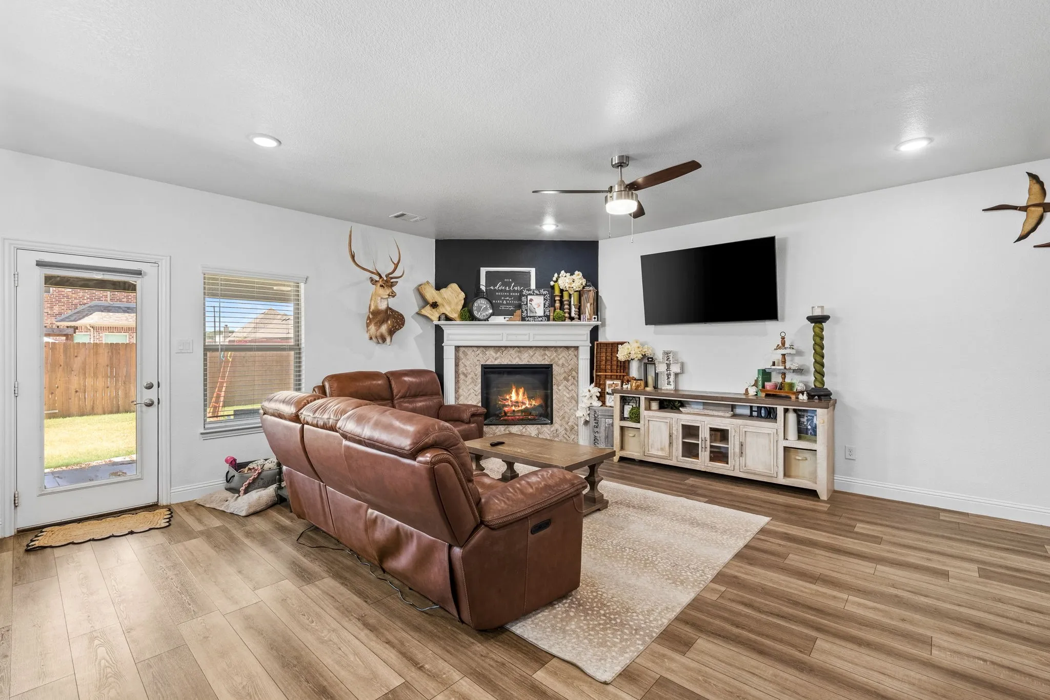 Wood-finish flooring throughout, ceiling fan, recessed lighting, corner fireplace with tile surround and mantel, and an exterior door leading to the backyard