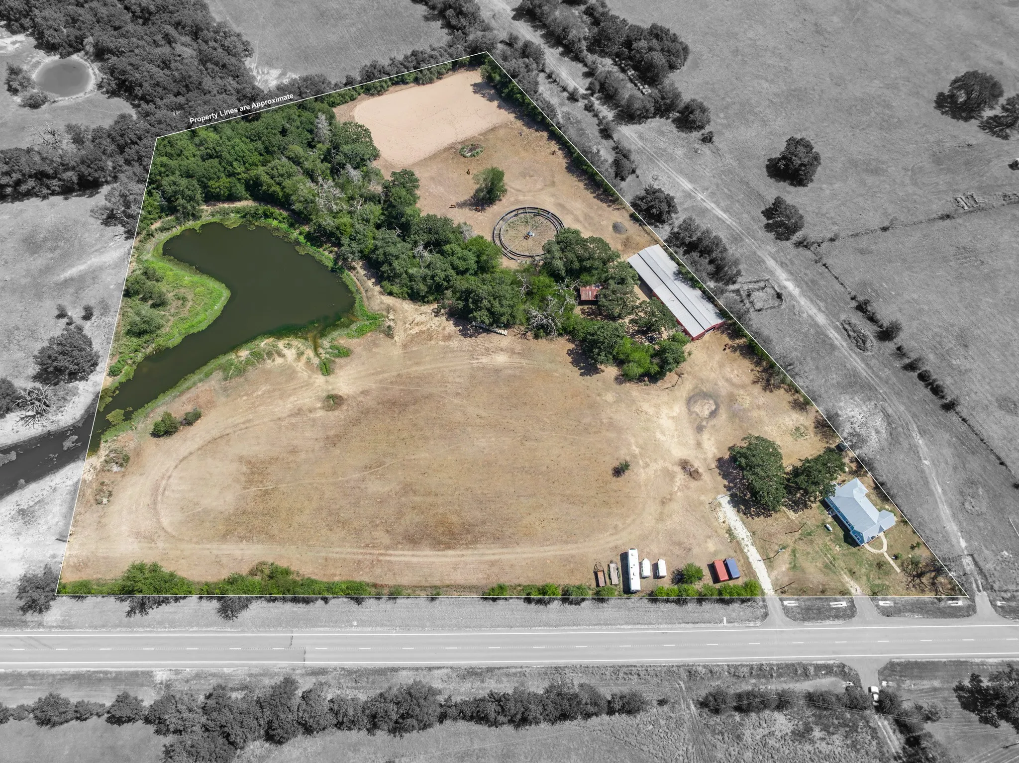 Expansive land featuring a large pond, a perimeter of mature trees, a residential structure, and a substantial outbuilding