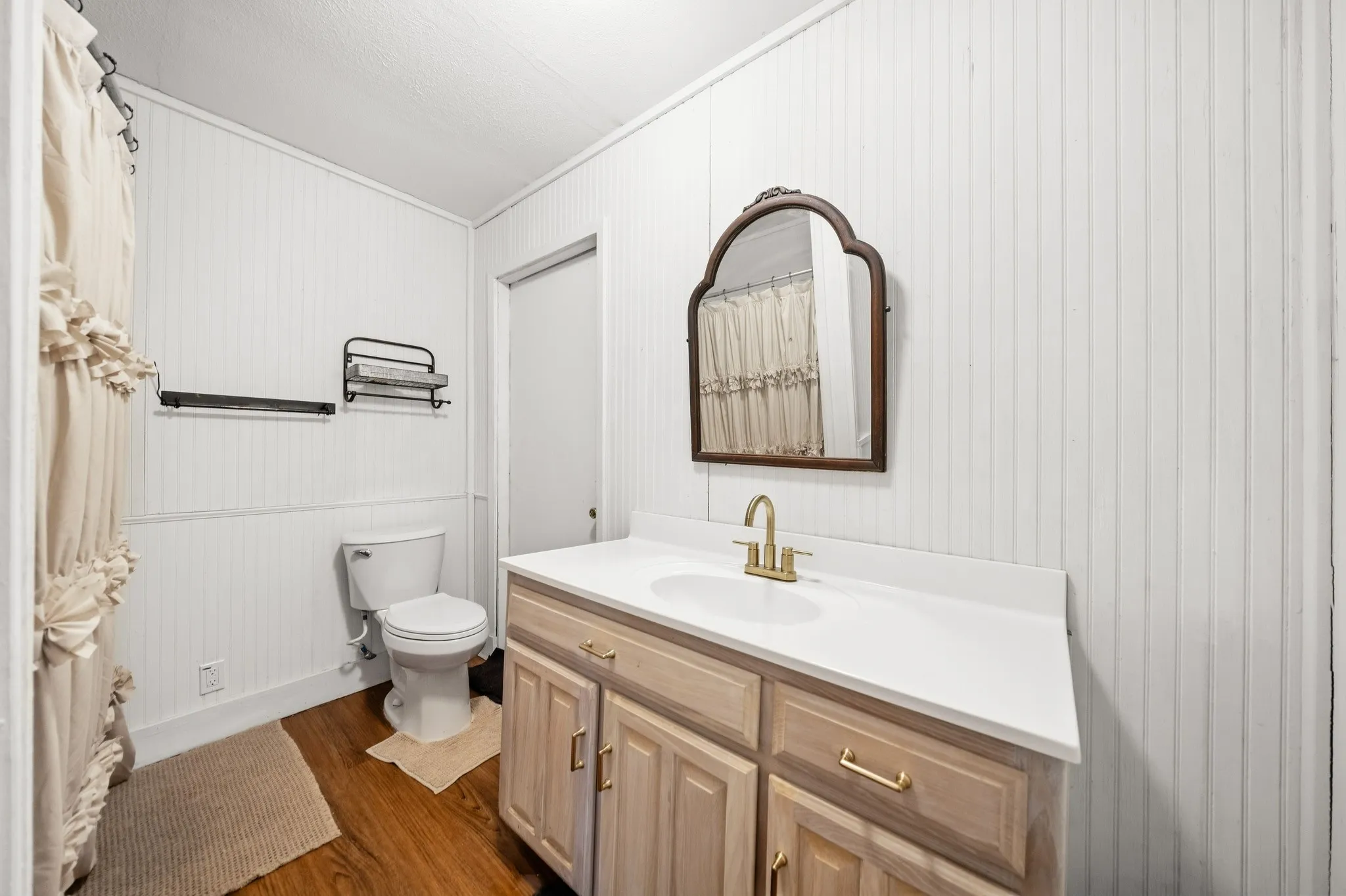 White beadboard walls, wood-finish flooring, light wood vanity with white countertop, gold-tone faucet, and an ornate framed mirror
