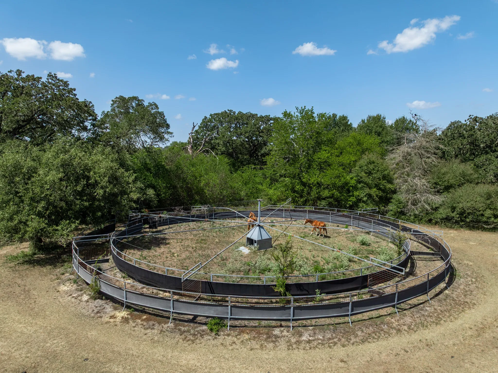 Circular fenced enclosure with a central covered structure, surrounded by mature trees and natural landscaping