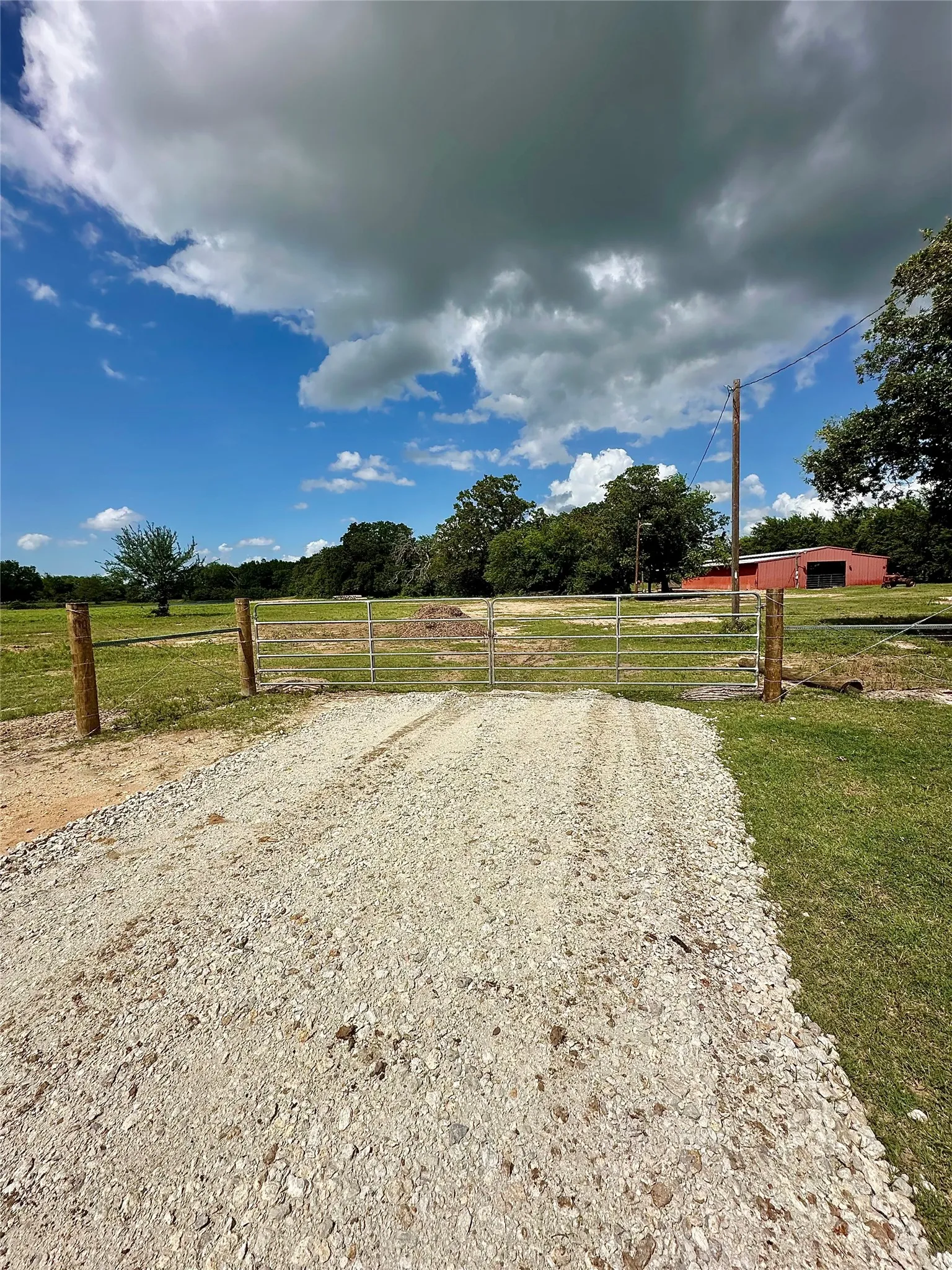 Gravel driveway with a metal gate and wooden fence posts, bordered by a grassy area and mature trees
