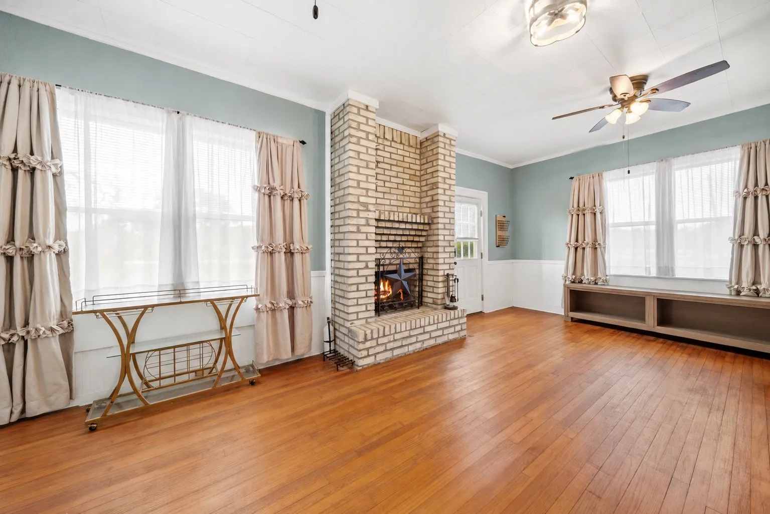 Wood-finish flooring, brick fireplace, built-in shelving, ceiling fan, and multiple windows