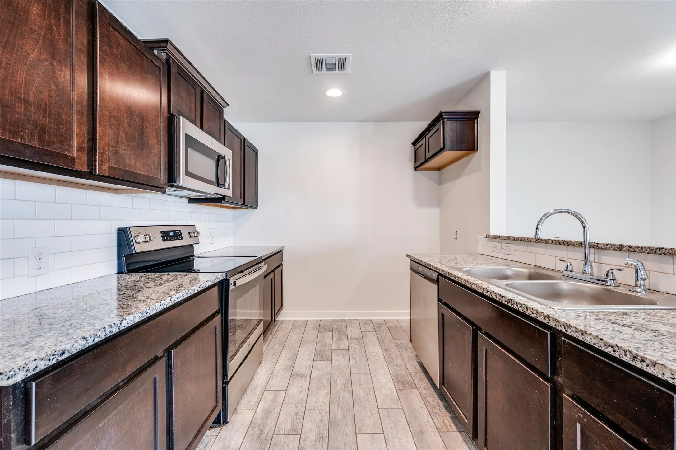 Kitchen featuring wood-finish flooring, dark wood cabinetry, granite countertops, a subway tile backsplash, and stainless steel appliances