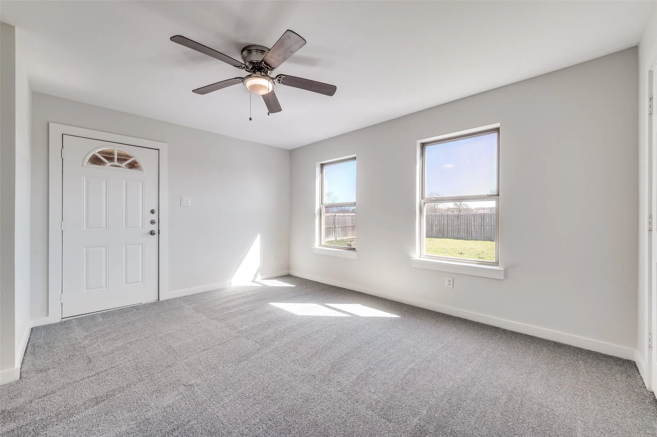 Spacious room featuring neutral carpet, white walls, two large windows, a ceiling fan with integrated lighting, and a paneled entry door with an arched glass insert