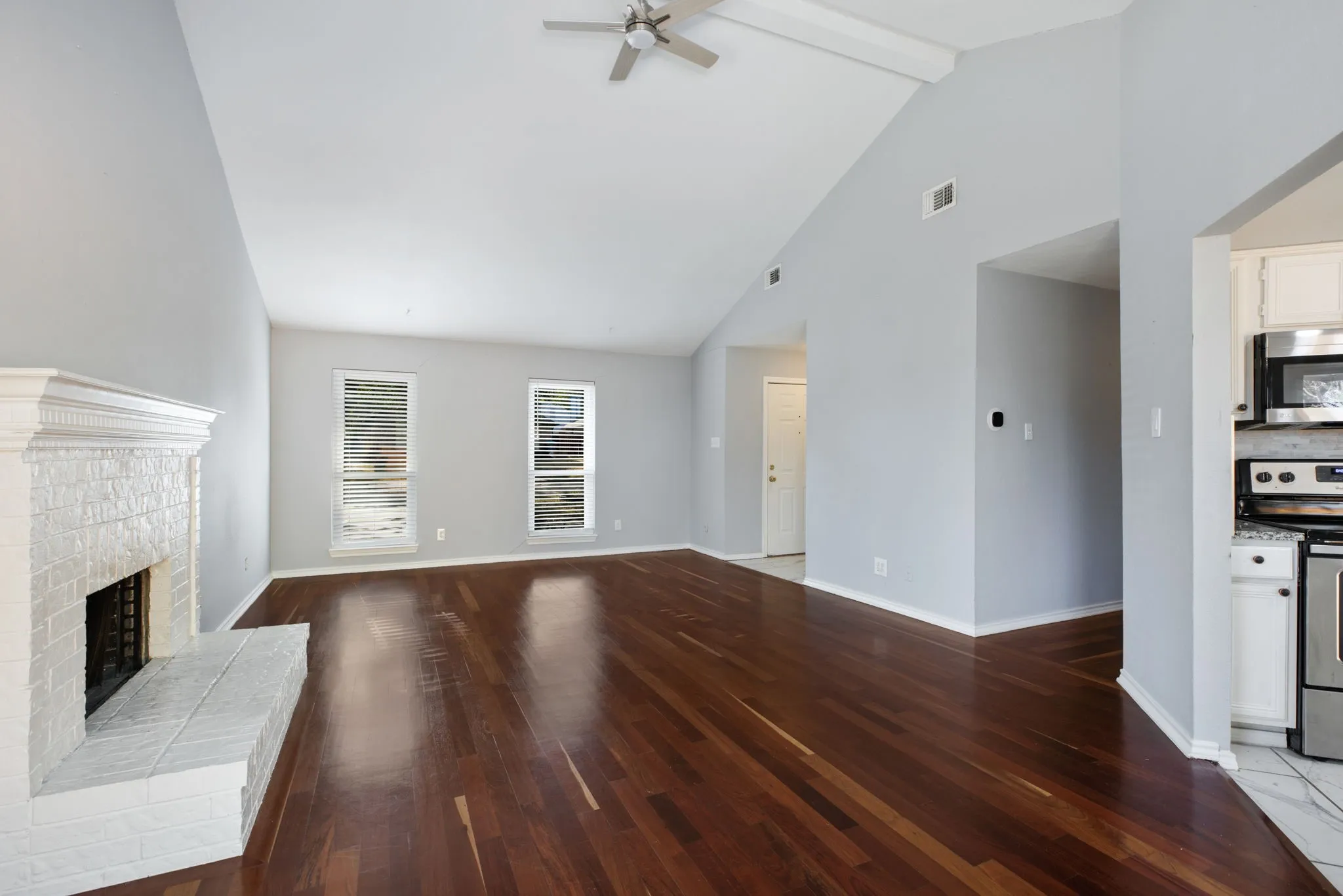 Vaulted ceiling with a ceiling fan, wood-finish flooring, white brick fireplace, dual windows, and an adjacent kitchen