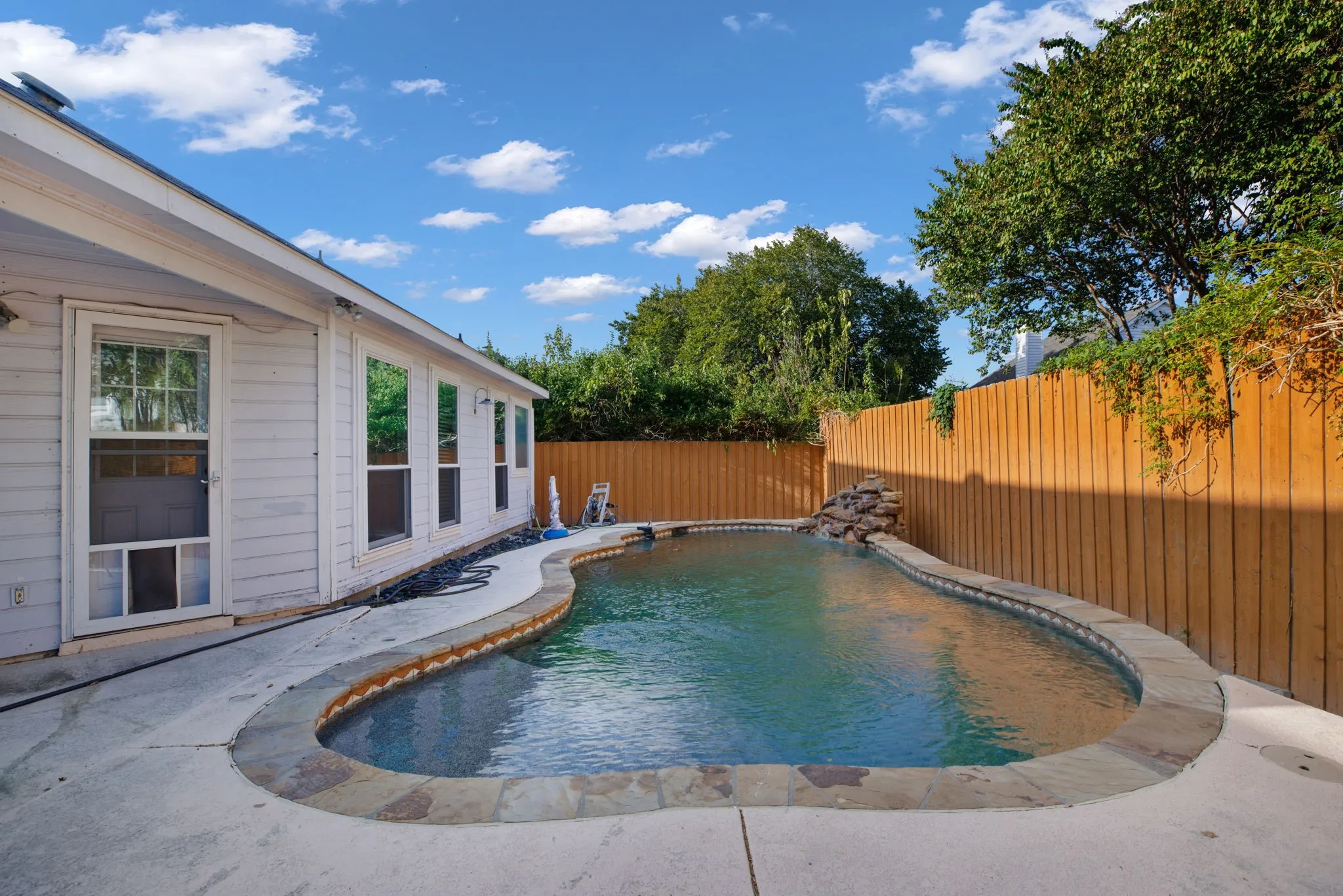 Curved swimming pool with stone coping, water feature, and surrounding concrete patio