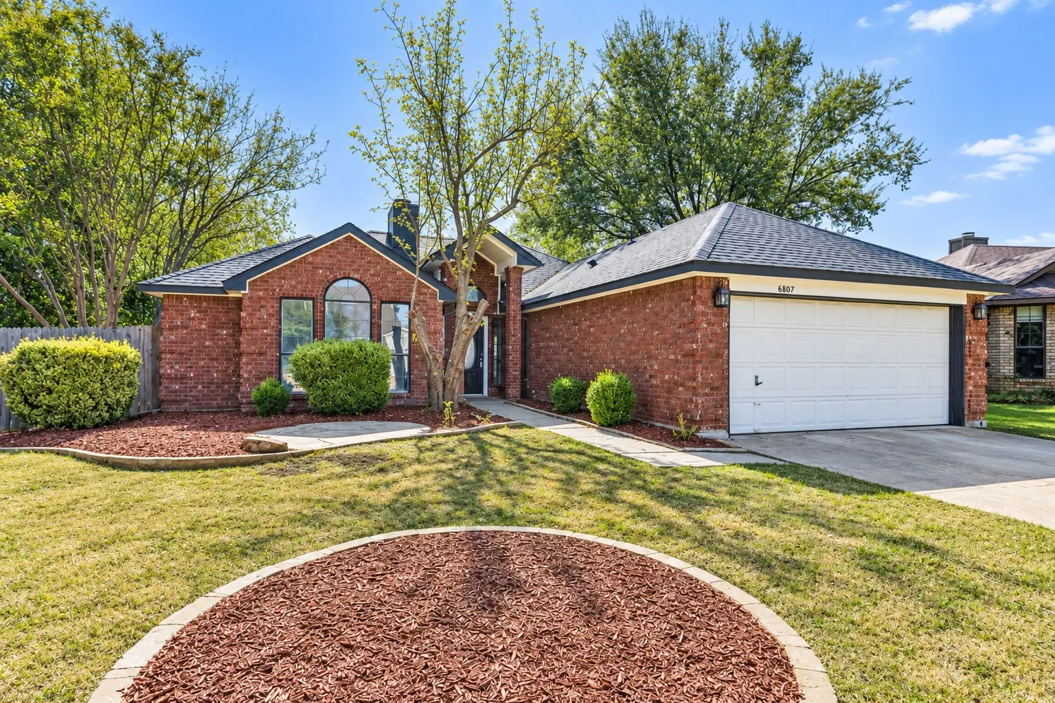 Brick exterior with a dark-shingled roof, arched window, attached garage, and landscaped front yard with mulch beds