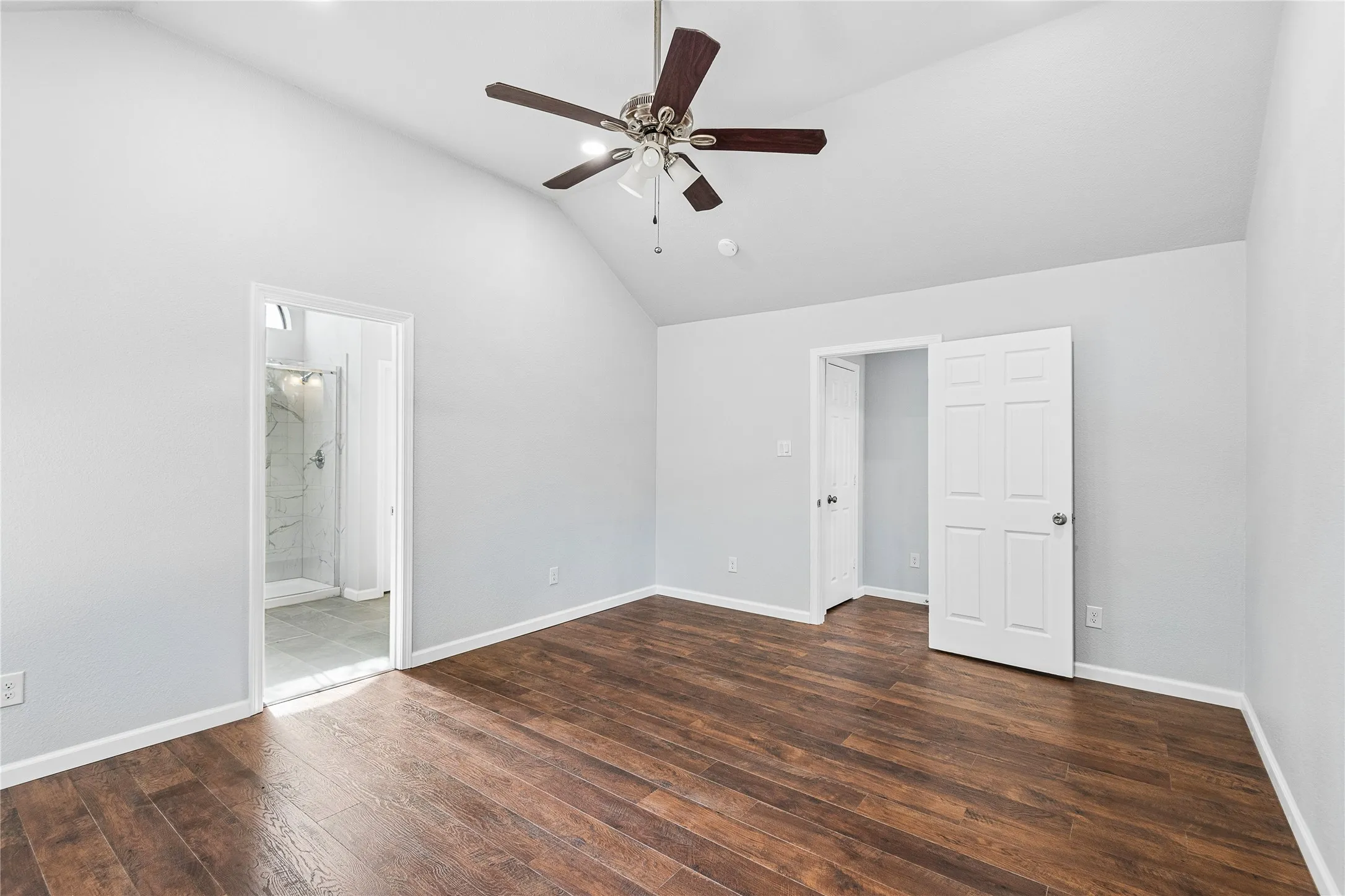Vaulted ceiling, wood-finish flooring, ceiling fan with light fixture, and an attached bathroom with a glass-enclosed shower