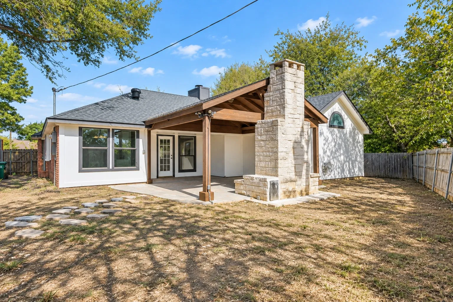 Covered patio with exposed wood beams, stone-clad outdoor fireplace, dual glass-panel patio doors, and a partial privacy fence