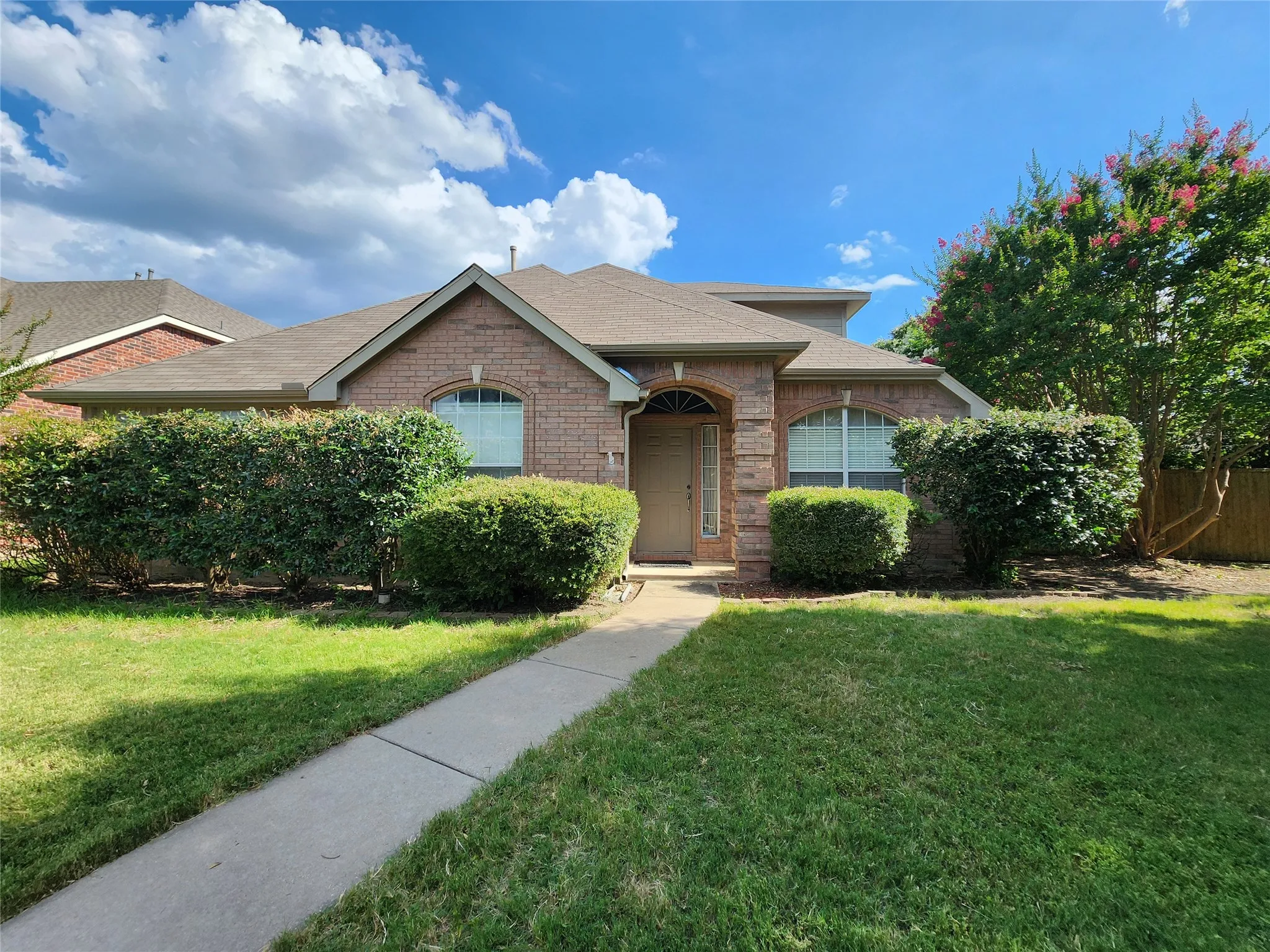 Brick exterior with gabled roofline, arched window, mature shrubs, and concrete walkway