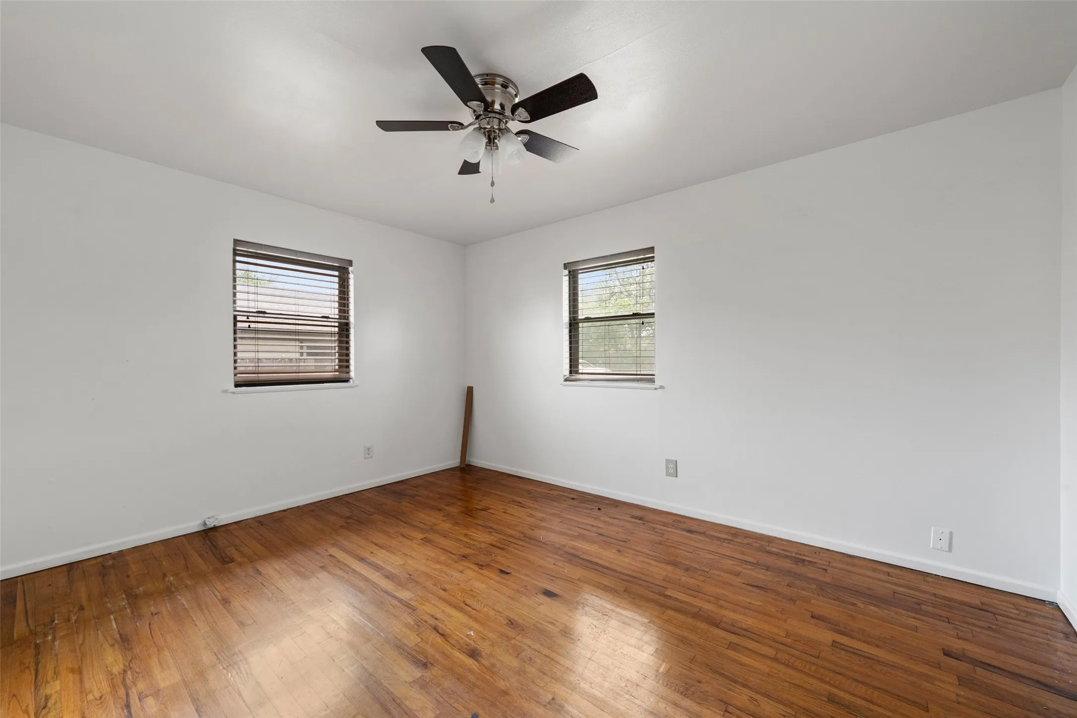 Bright corner bedroom showcasing rich wood-finish flooring, ceiling fan with integrated lighting, and dual windows with horizontal blinds providing natural light and ventilation control