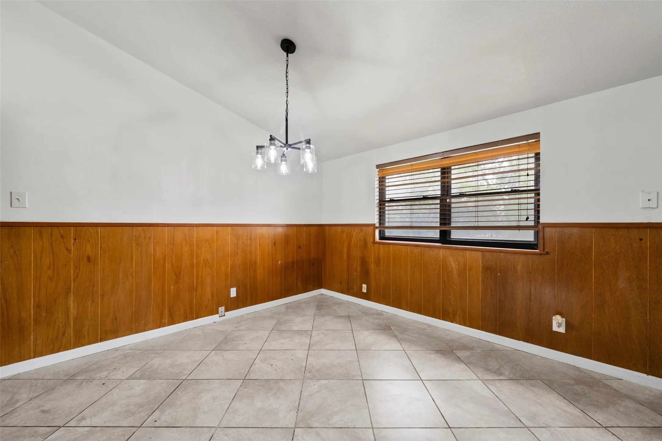Vaulted ceiling with an updated chandelier, wood-paneled wainscoting, and tile flooring