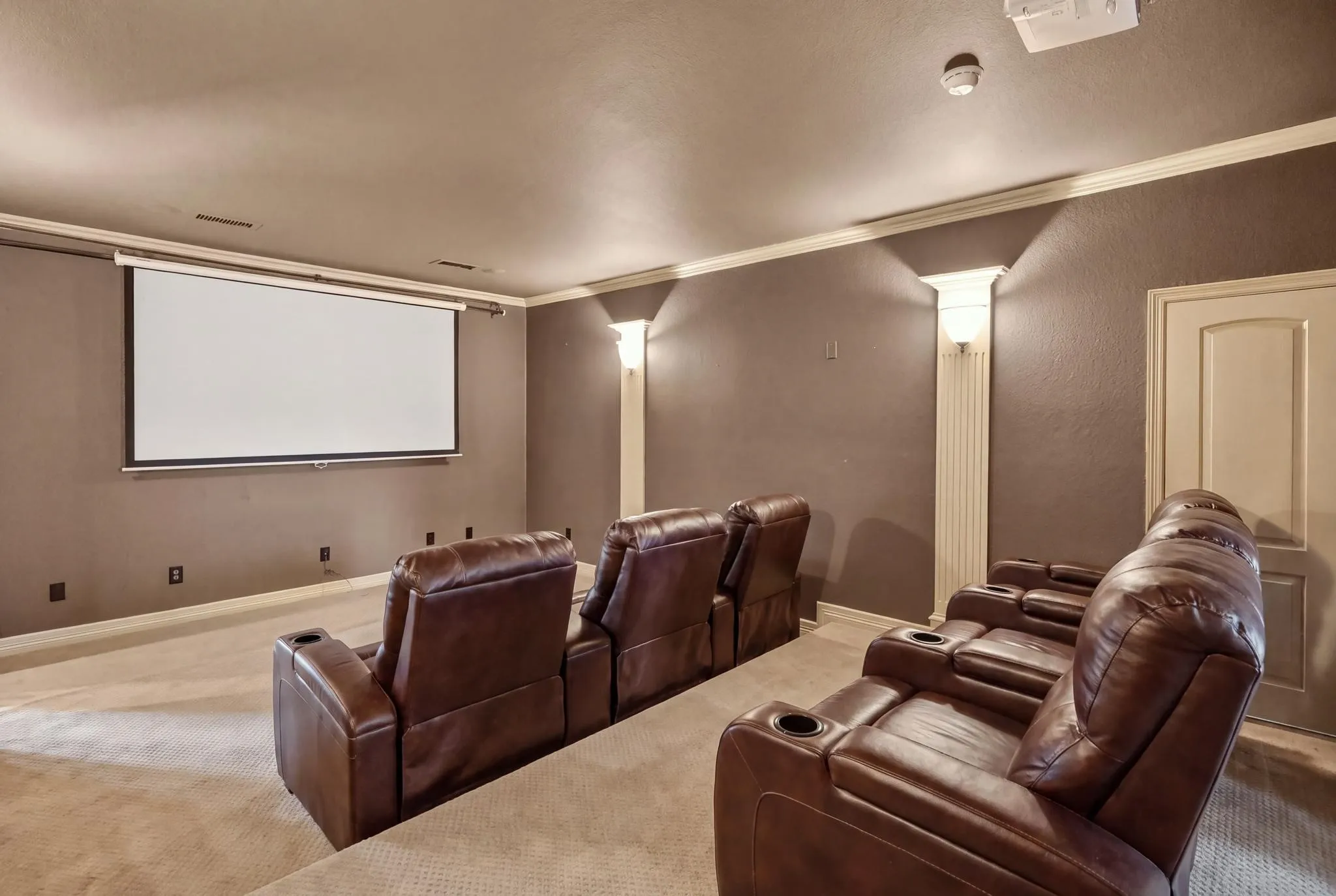 Dedicated two tier media room with a projector screen, wall sconces, ceiling crown molding, and light-colored carpeting. Virtually Staged