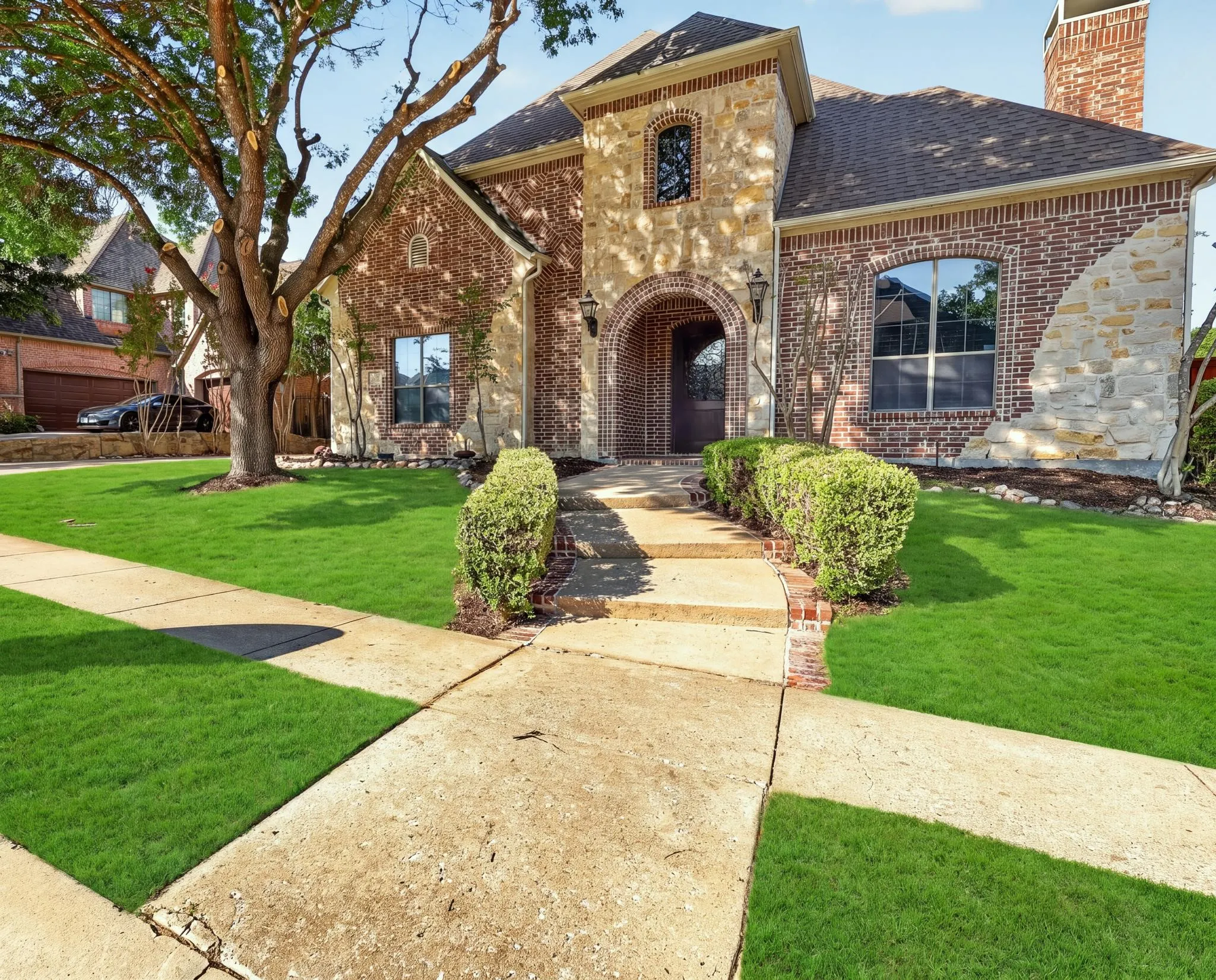 Brick and stone exterior with an arched entryway, mature front tree, and a paved walkway bordered by manicured shrubs