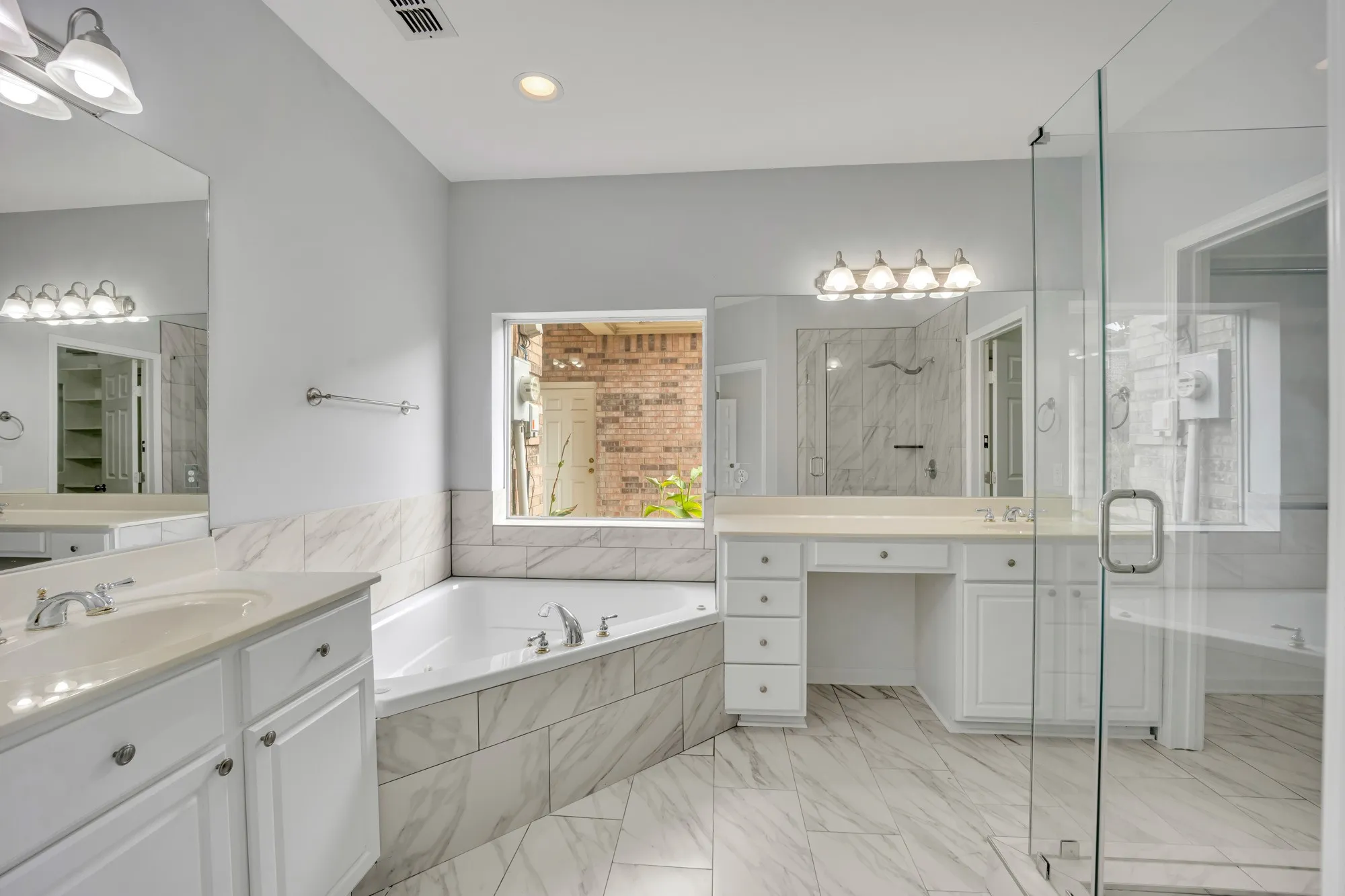 Spacious bathroom featuring a built-in corner tub, glass-enclosed shower, dual vanities with white cabinetry, and large format tile flooring