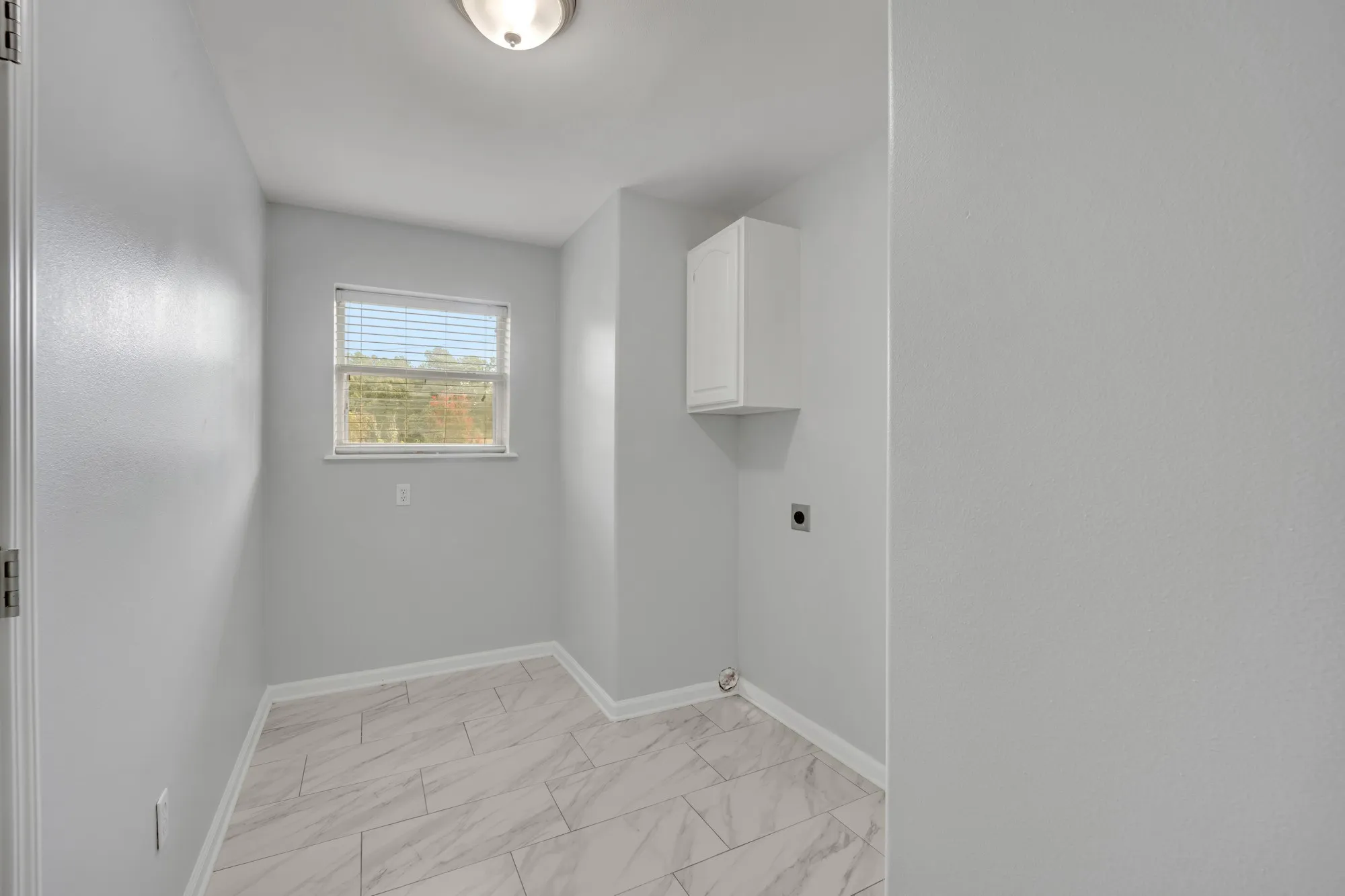 Window with horizontal blinds, overhead flush mount light fixture, wall-mounted white cabinetry, and large format floor tiling