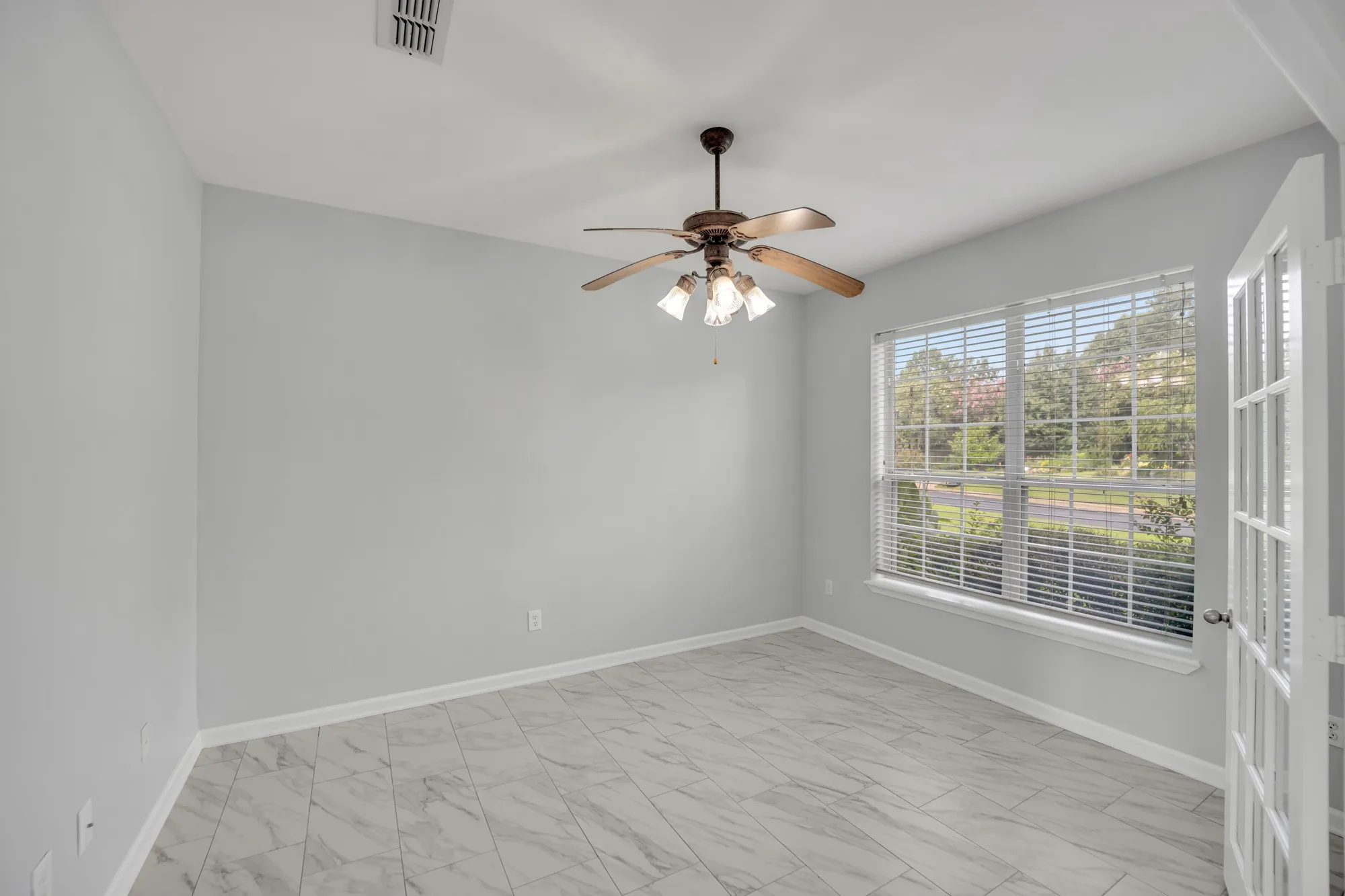 Wood-finish ceiling fan with integrated lighting, large window with blinds, white trim, light gray walls, and light-toned tile flooring